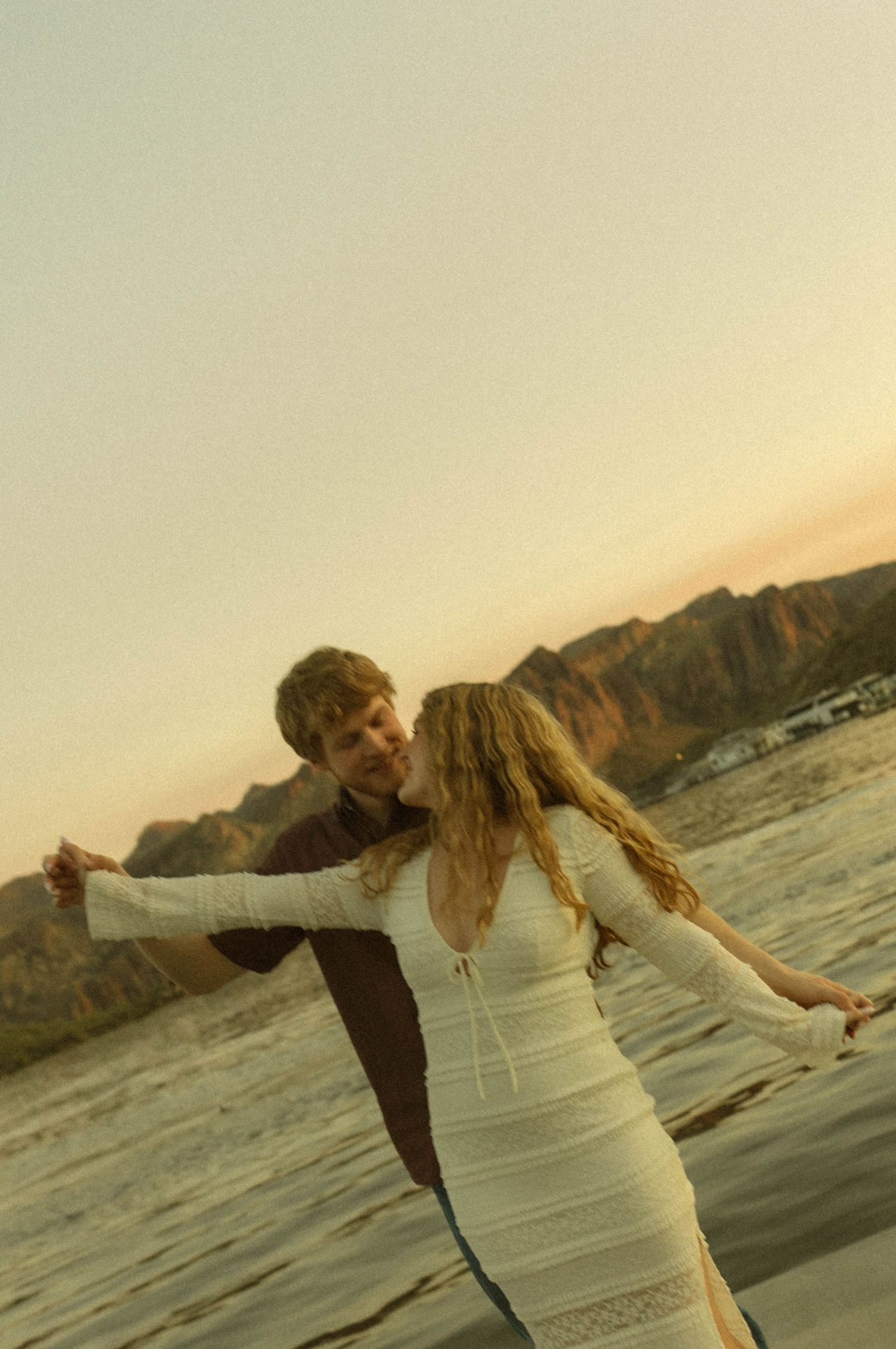 A couple in white clothing dances together on a beach at sunset with mountains visible in the background.