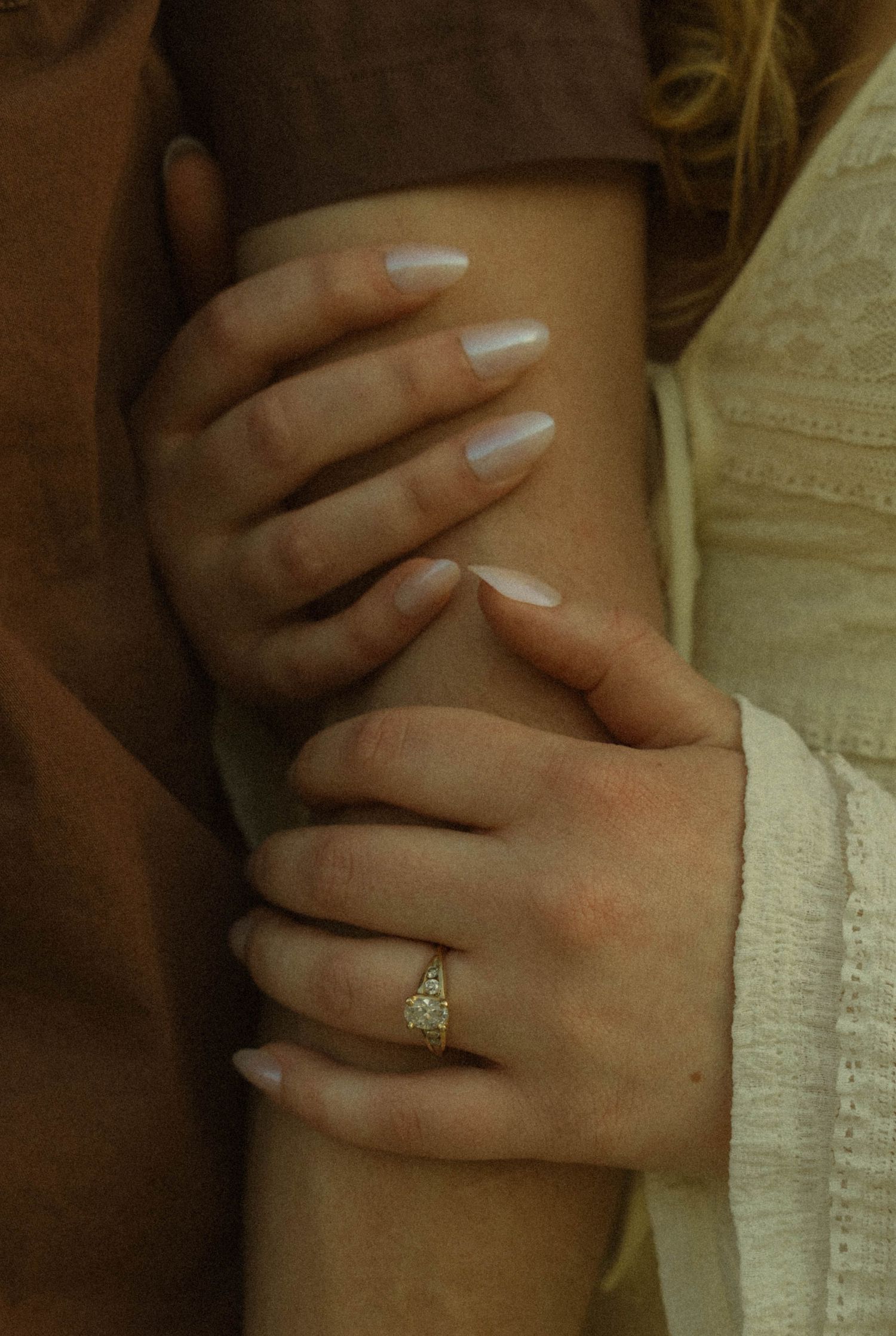 Close-up of hands embracing with a diamond ring, showing white manicured nails against cream-colored clothing.