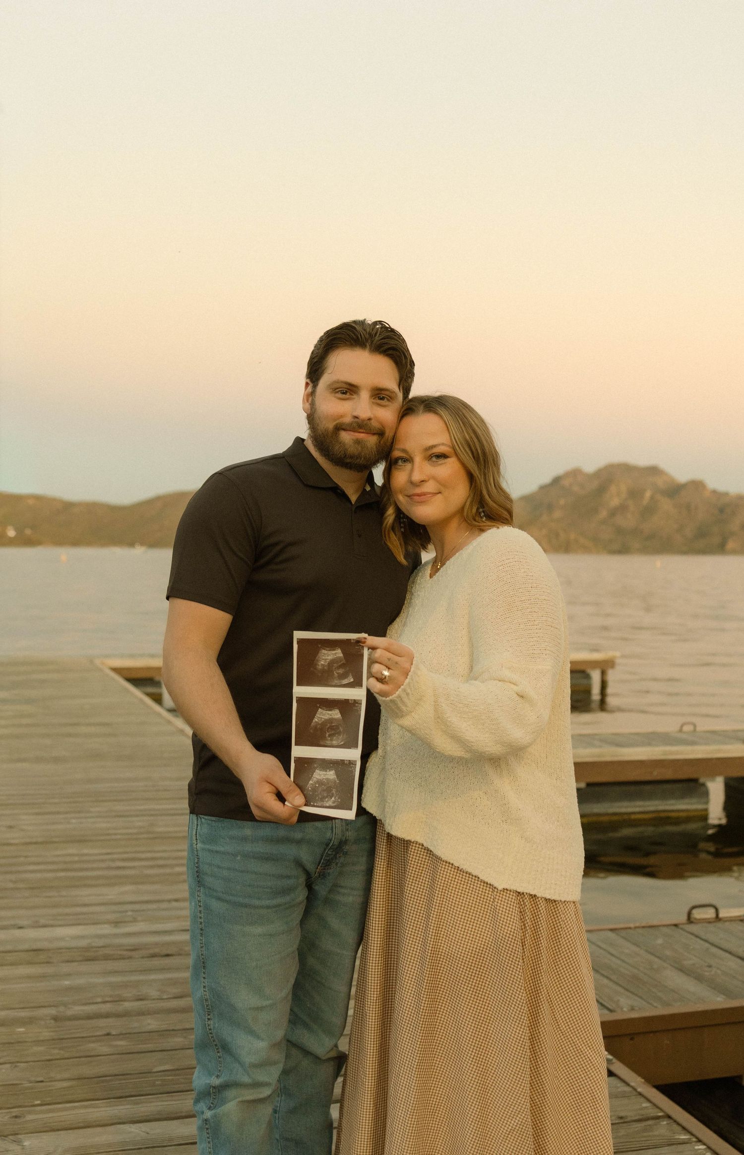 A couple stands on a dock at sunset holding an ultrasound photo while embracing.