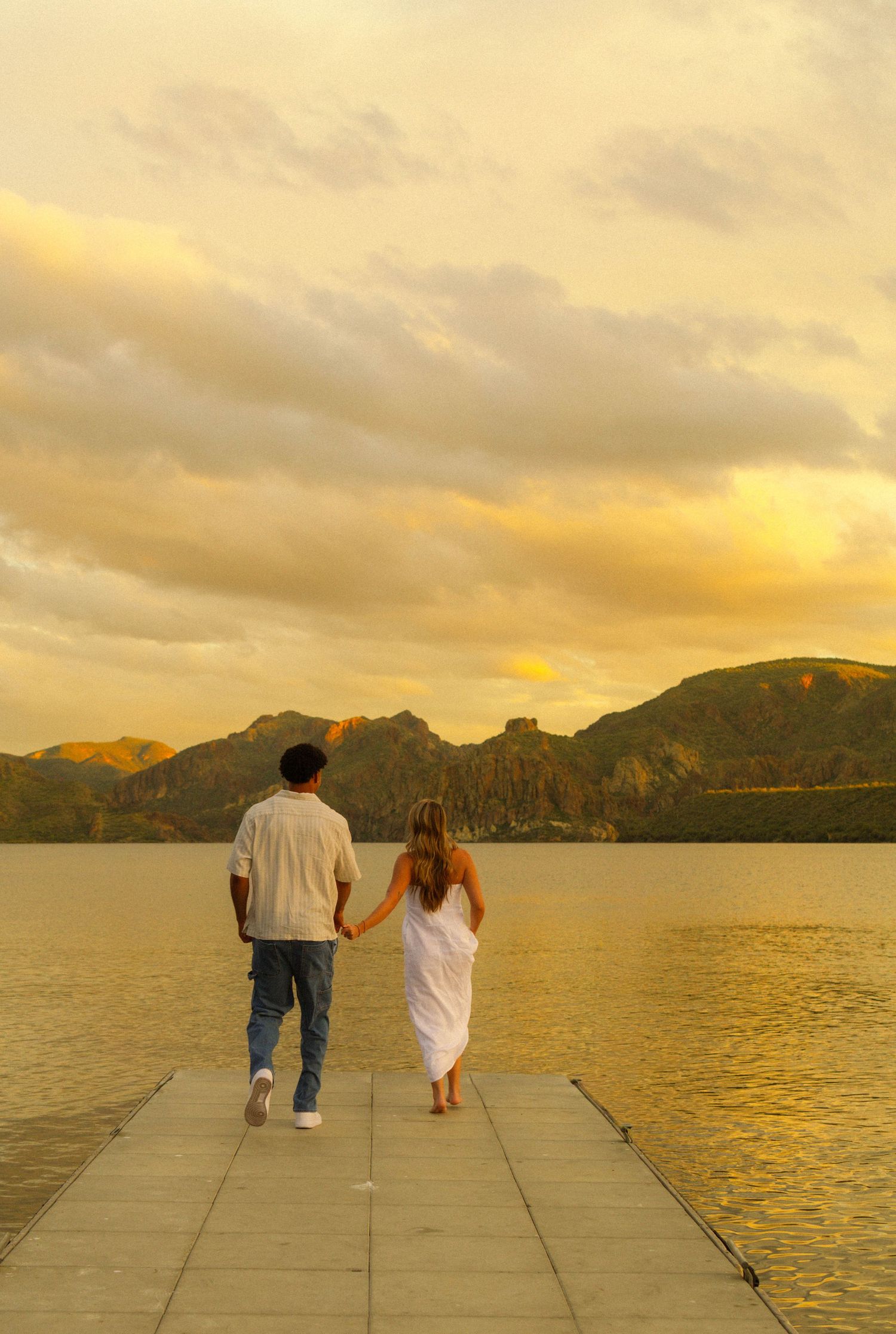 A couple stands on a pier overlooking a lake at sunset with mountains in the background under golden skies.