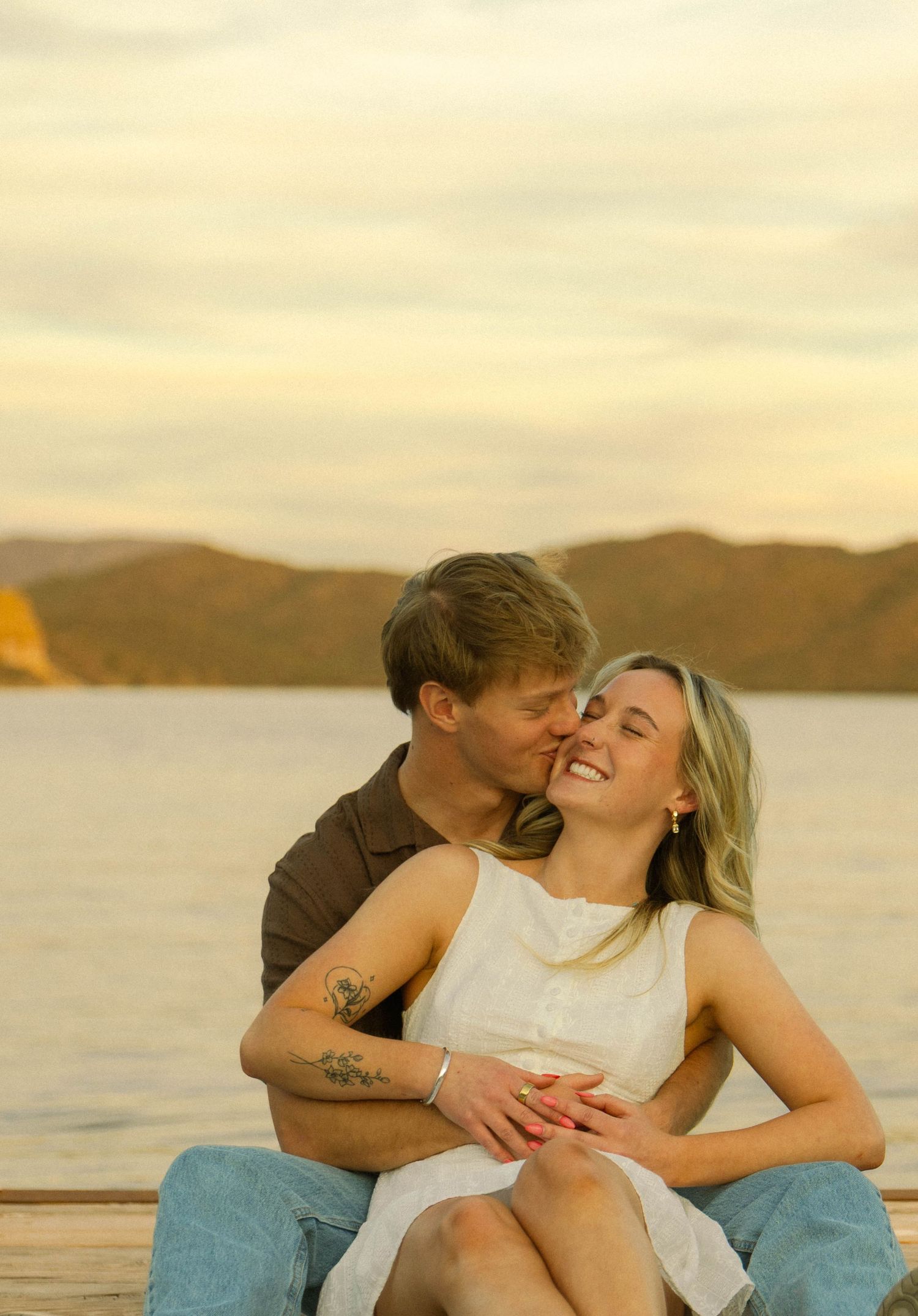 A couple embraces lovingly on a dock during golden hour with a serene lake and mountains behind them.