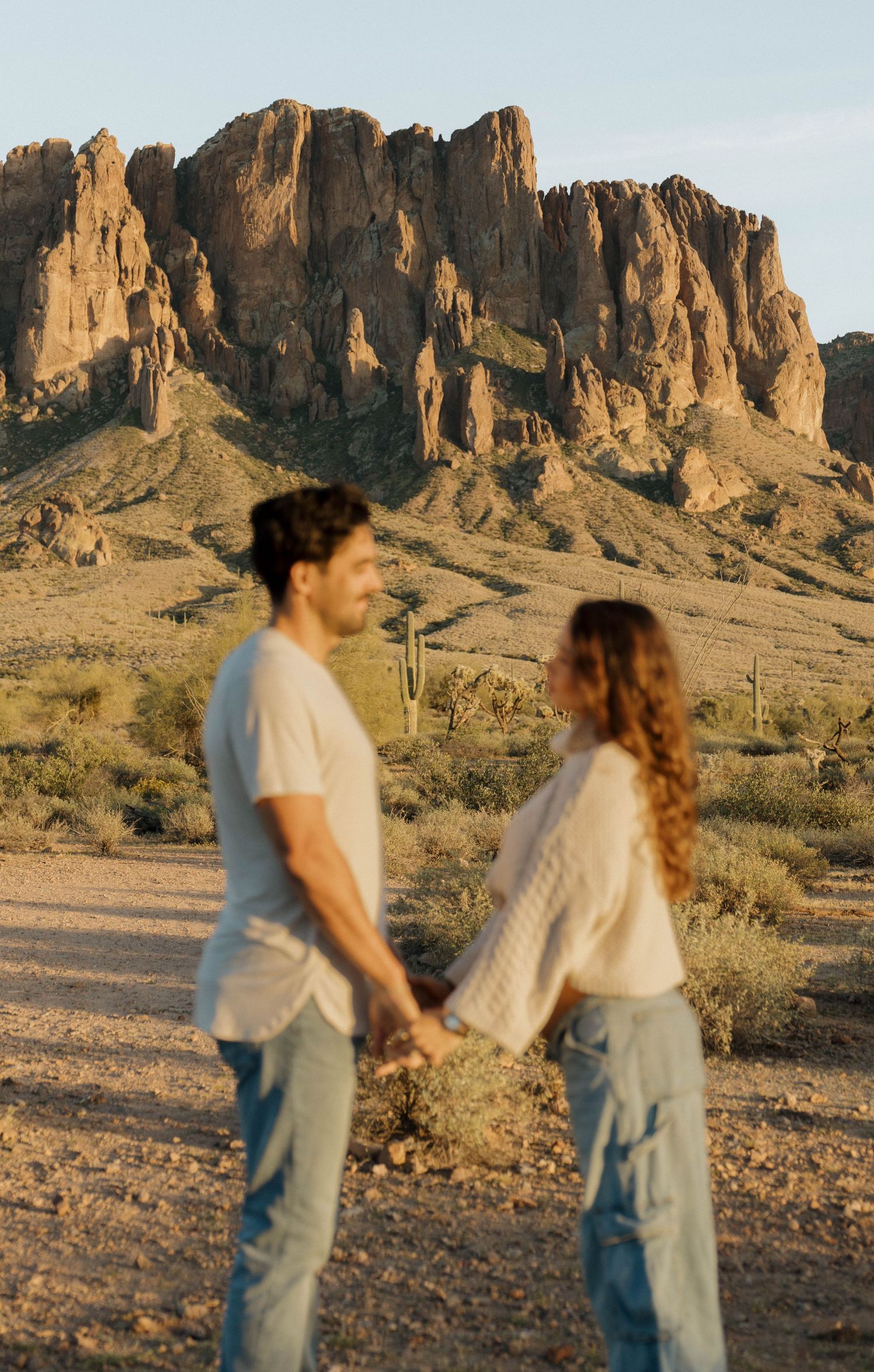 A couple holds hands while standing in front of the majestic Superstition Mountains in Arizona at golden hour.