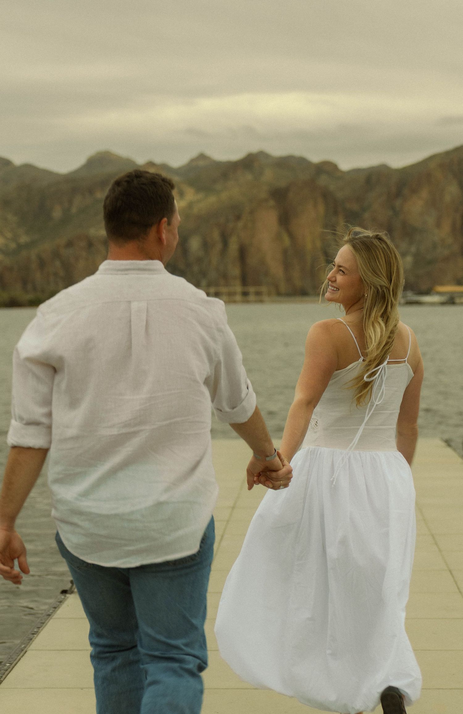 A couple in matching white outfits walks hand in hand along a lakeside dock with mountains behind them.