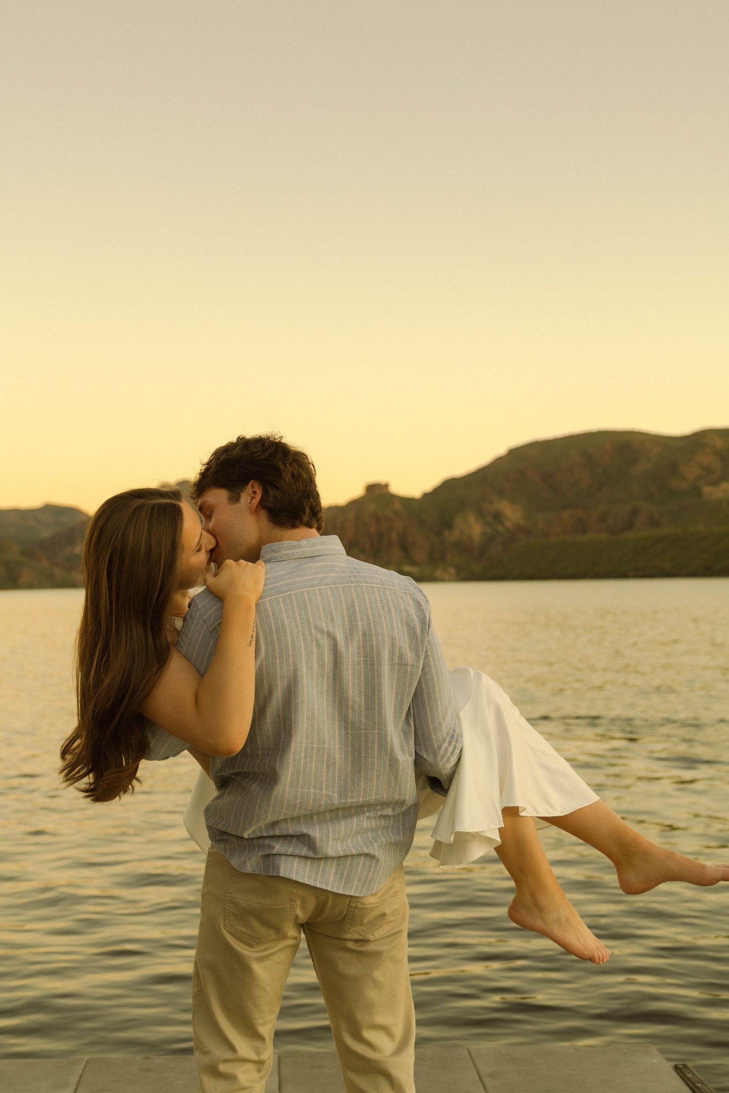 A couple in light colored clothing shares a tender moment by a lake during sunset.
