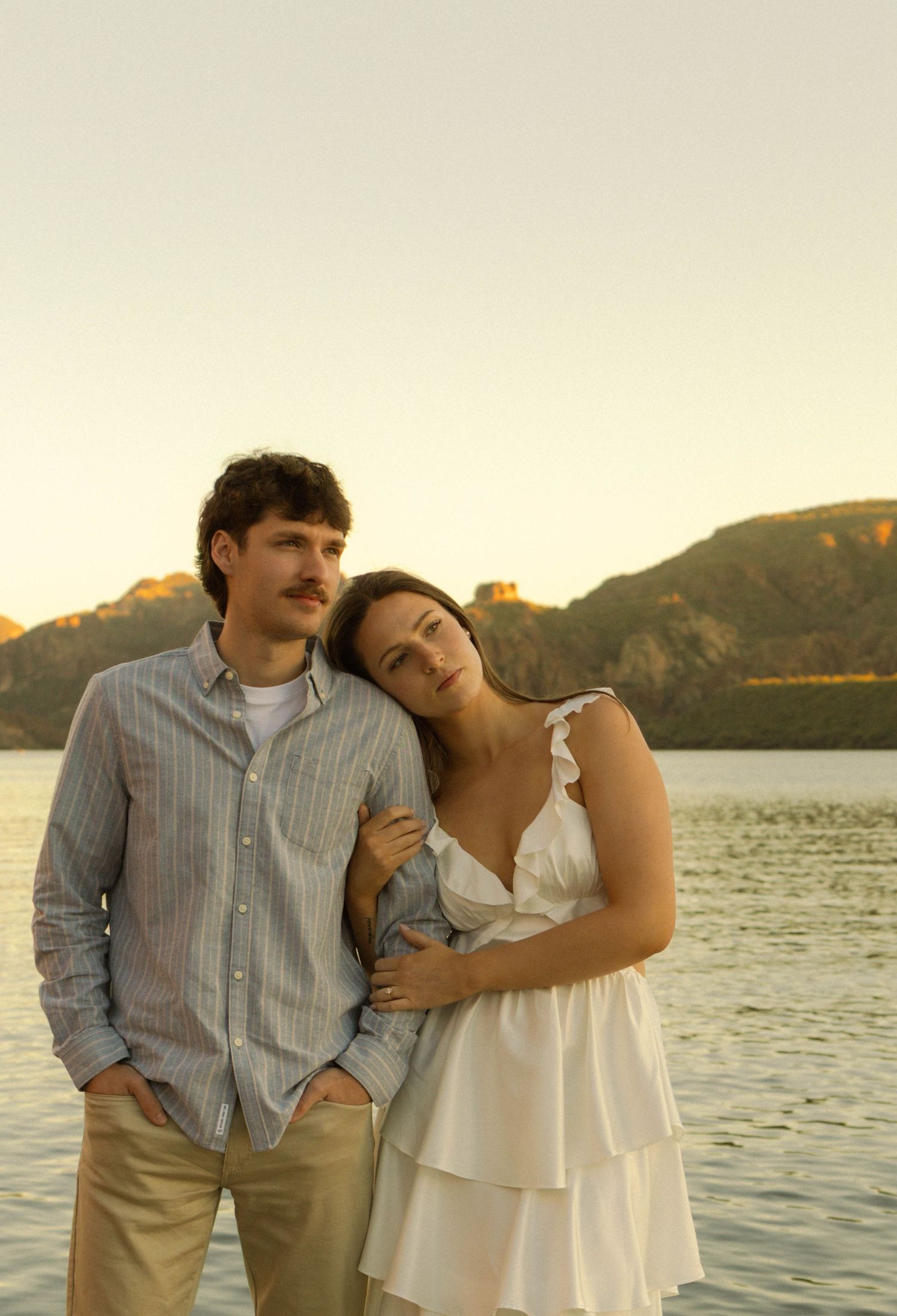 A couple embraces by the water during golden hour with mountains in the background.