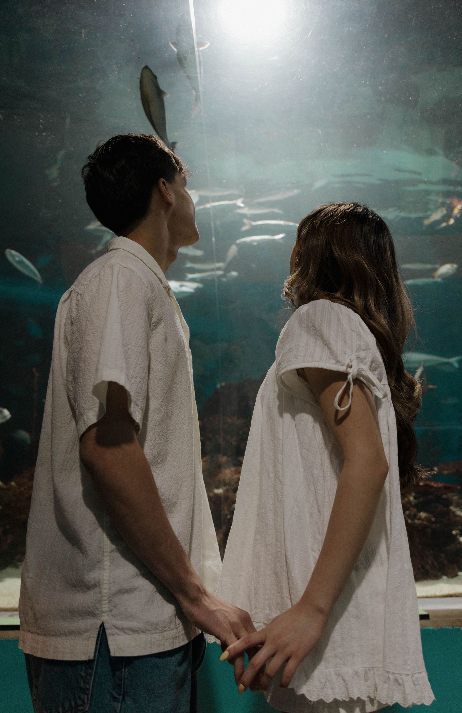 A couple in white clothing holds hands while watching fish swim in an aquarium display.