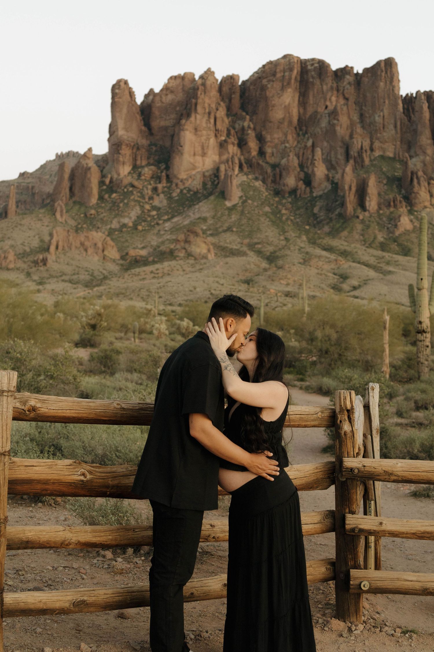 A romantic couple's photoshoot at sunset by a wooden fence with desert mountains in the background.