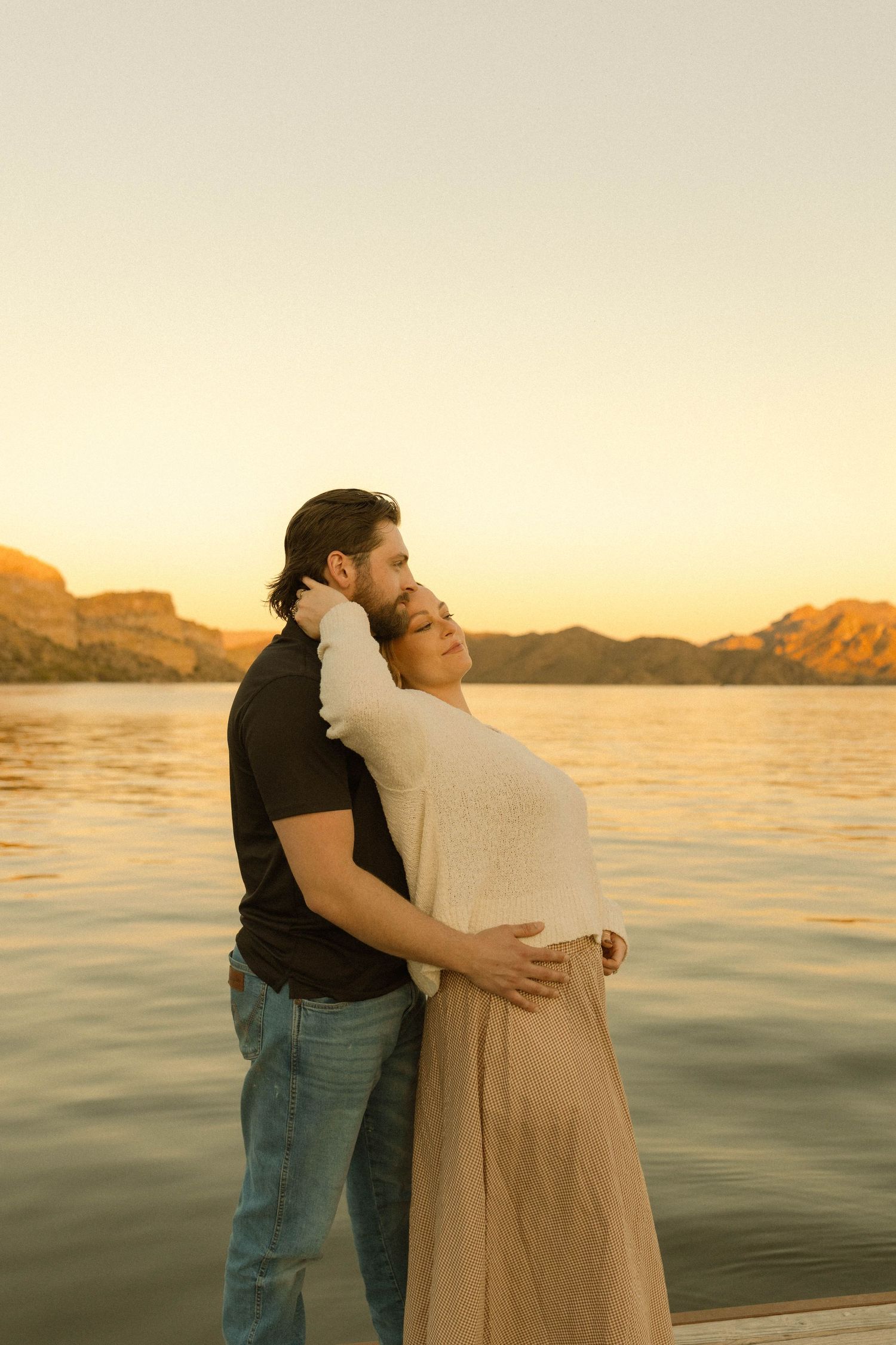 Intimate moment between couple standing by waterfront during golden hour.
