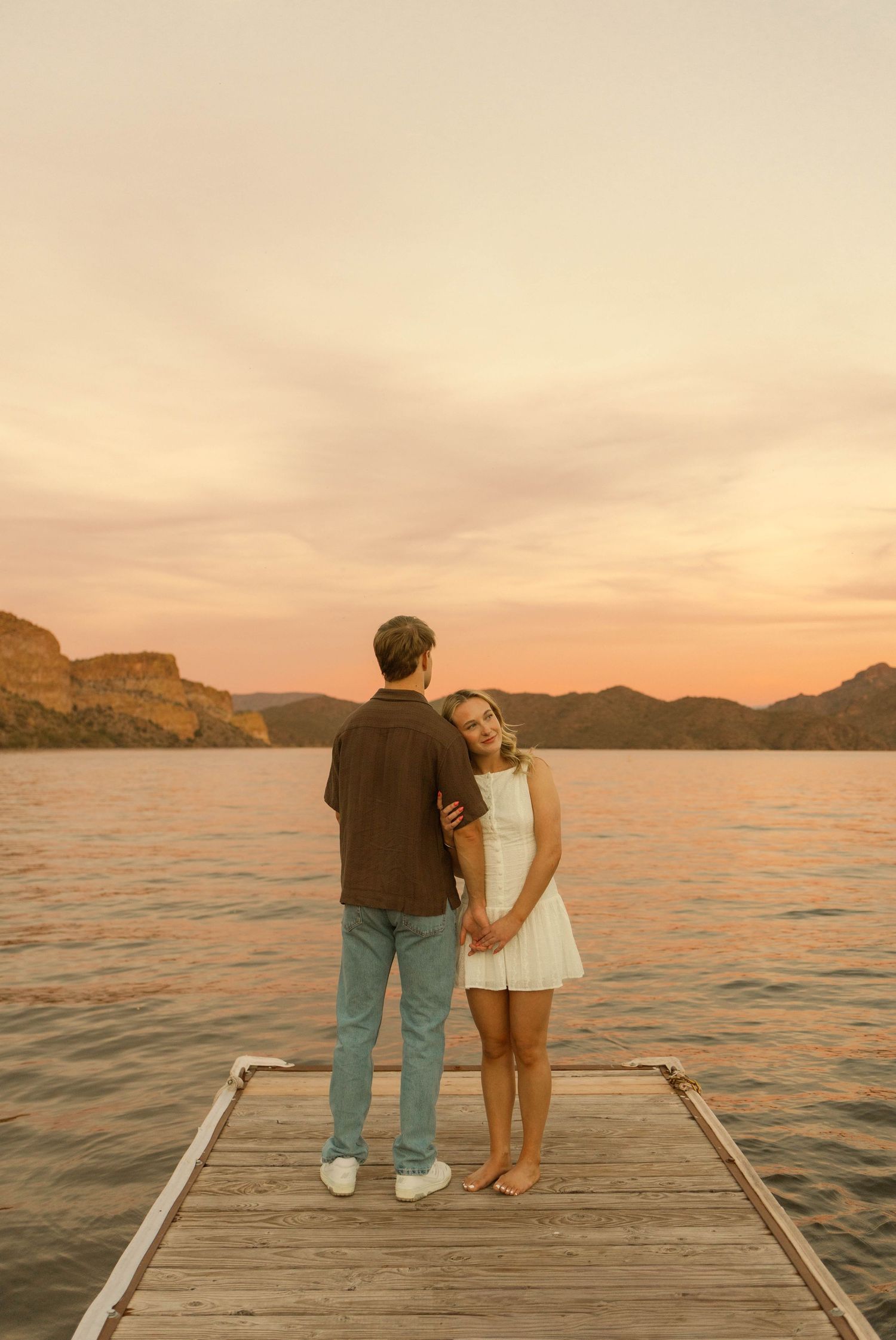 A couple shares a romantic moment on a wooden dock at sunset over a serene lake with mountains in the background.