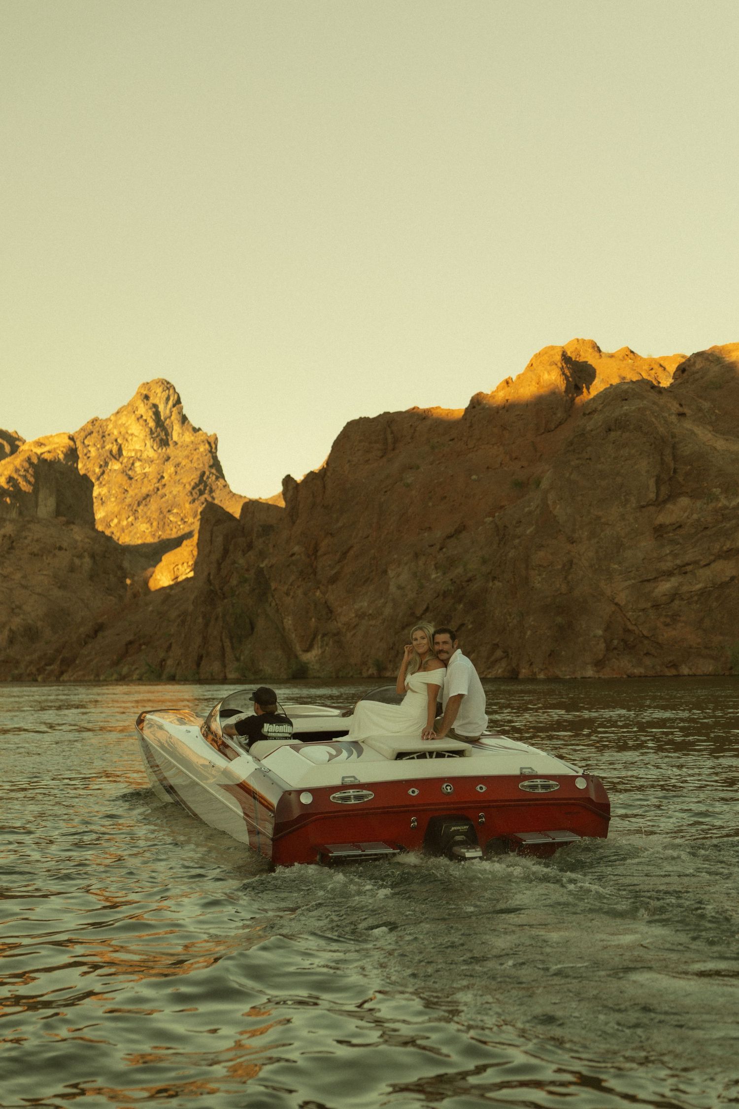 Couple on a red and white speedboat cruising on calm waters with dramatic rocky mountains at golden sunset.
