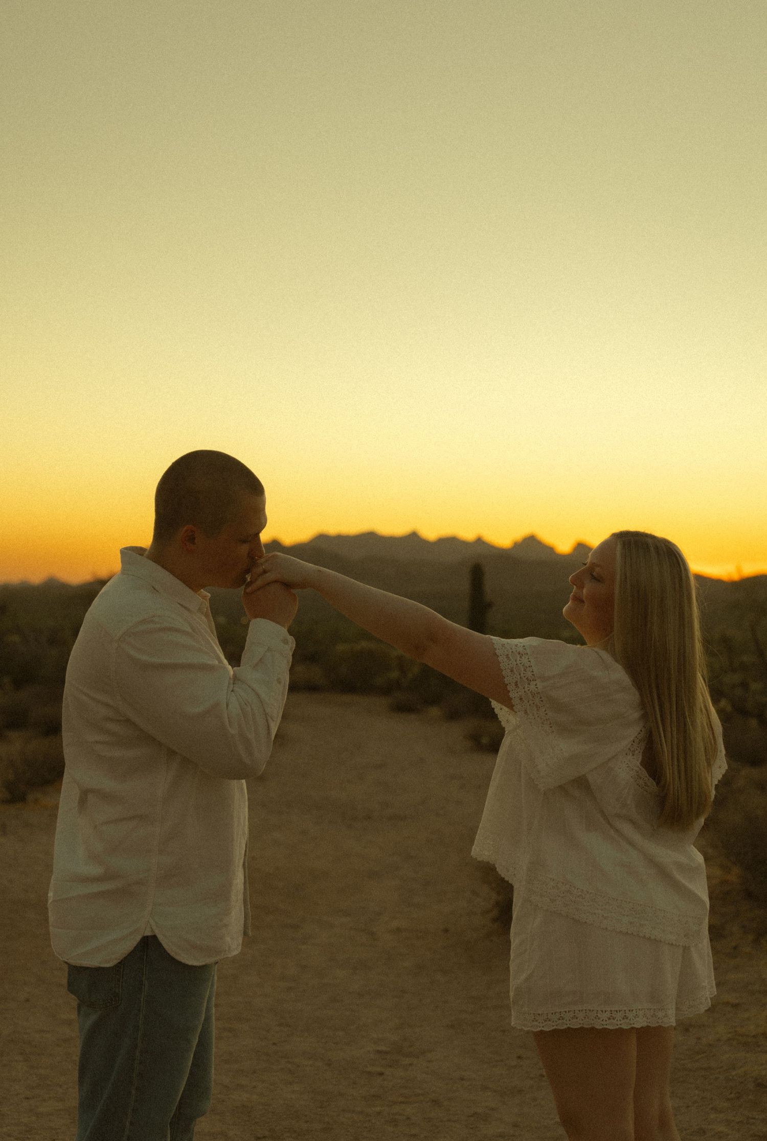 A couple shares a romantic moment at sunset in the desert, man kissing woman's hand in golden light.