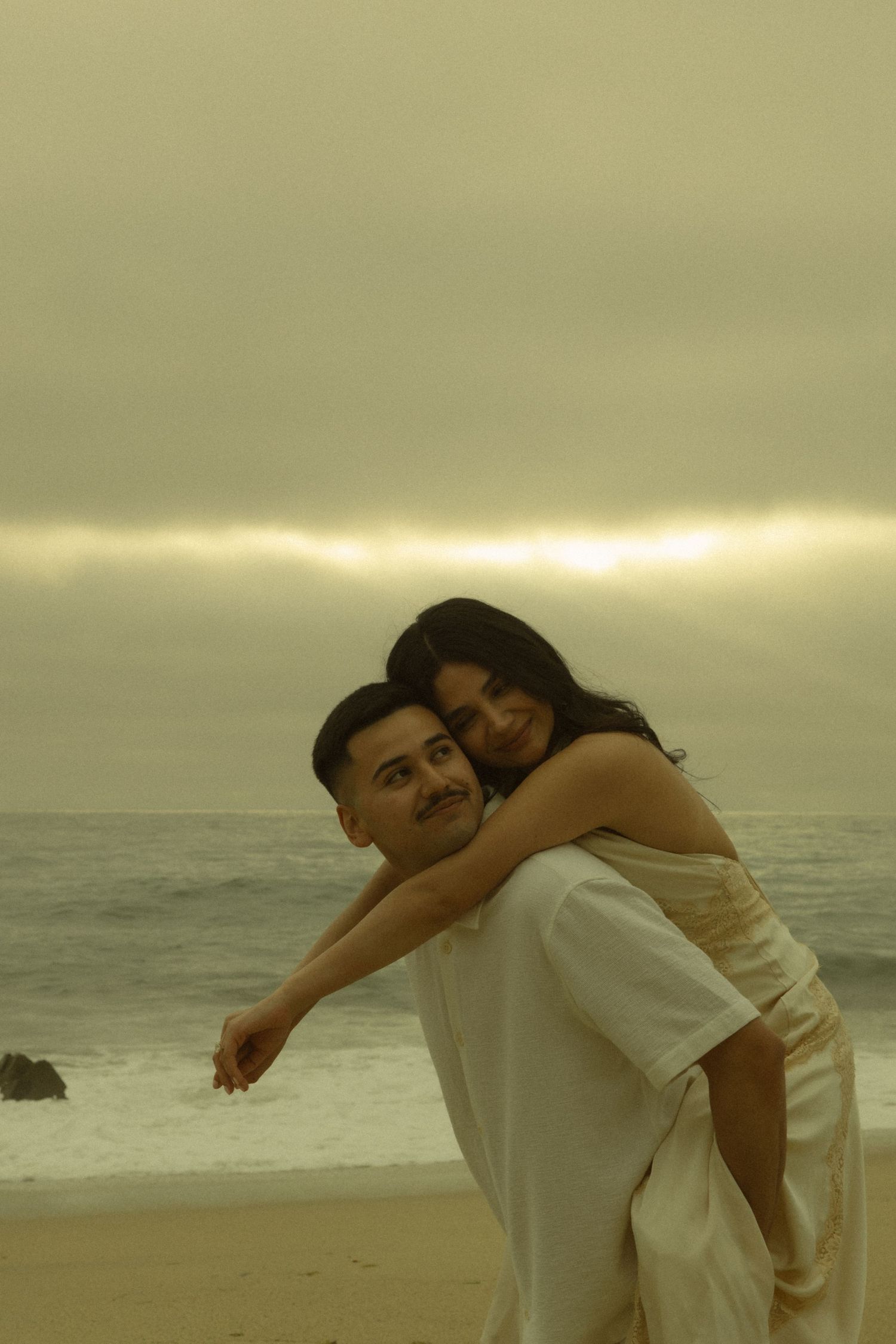 Happy couple on beach, man giving woman a piggyback ride, smiling with ocean and sunset behind them.