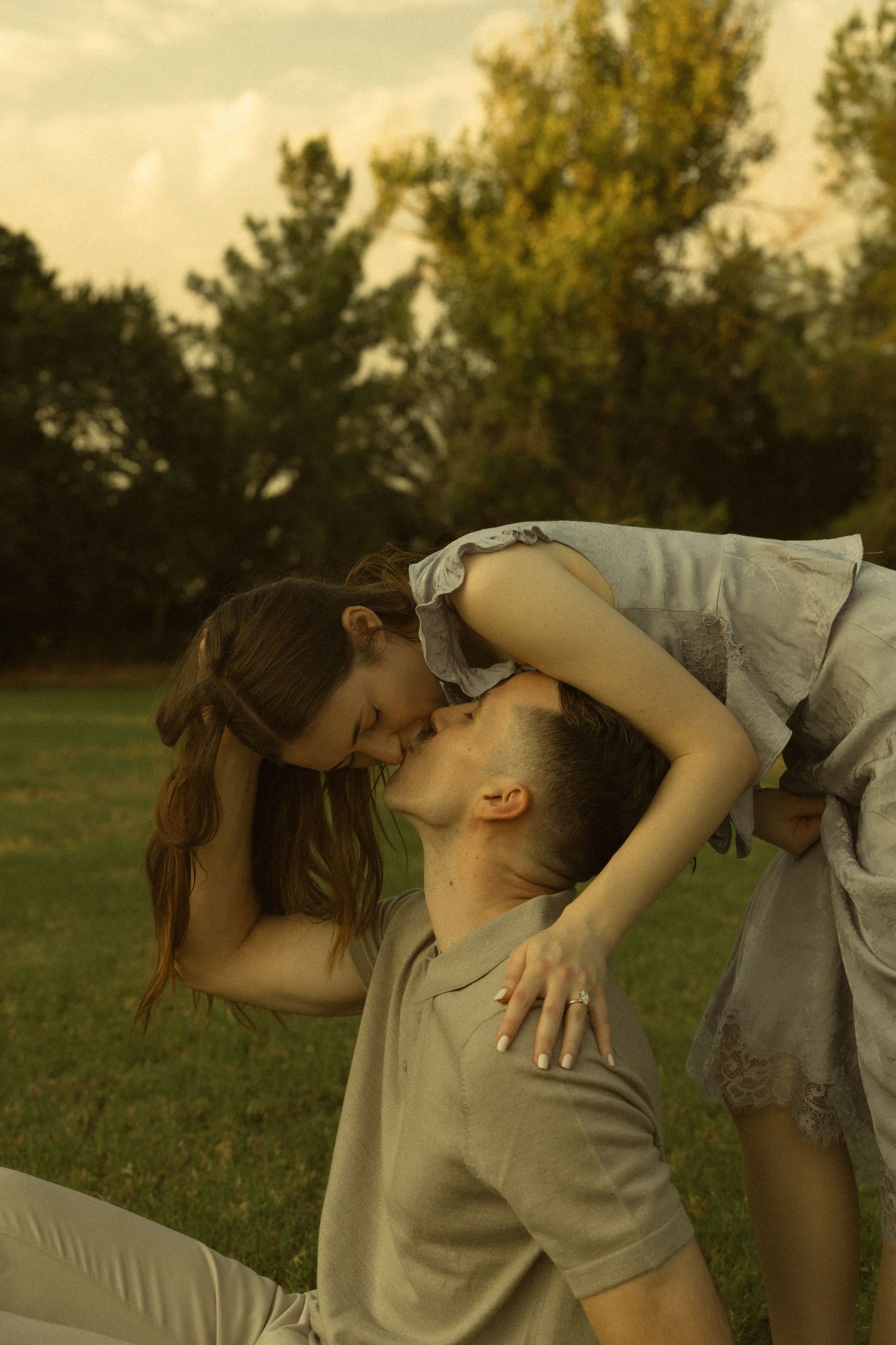Romantic couple sharing a tender kiss outdoors on green grass with trees in the background at sunset.