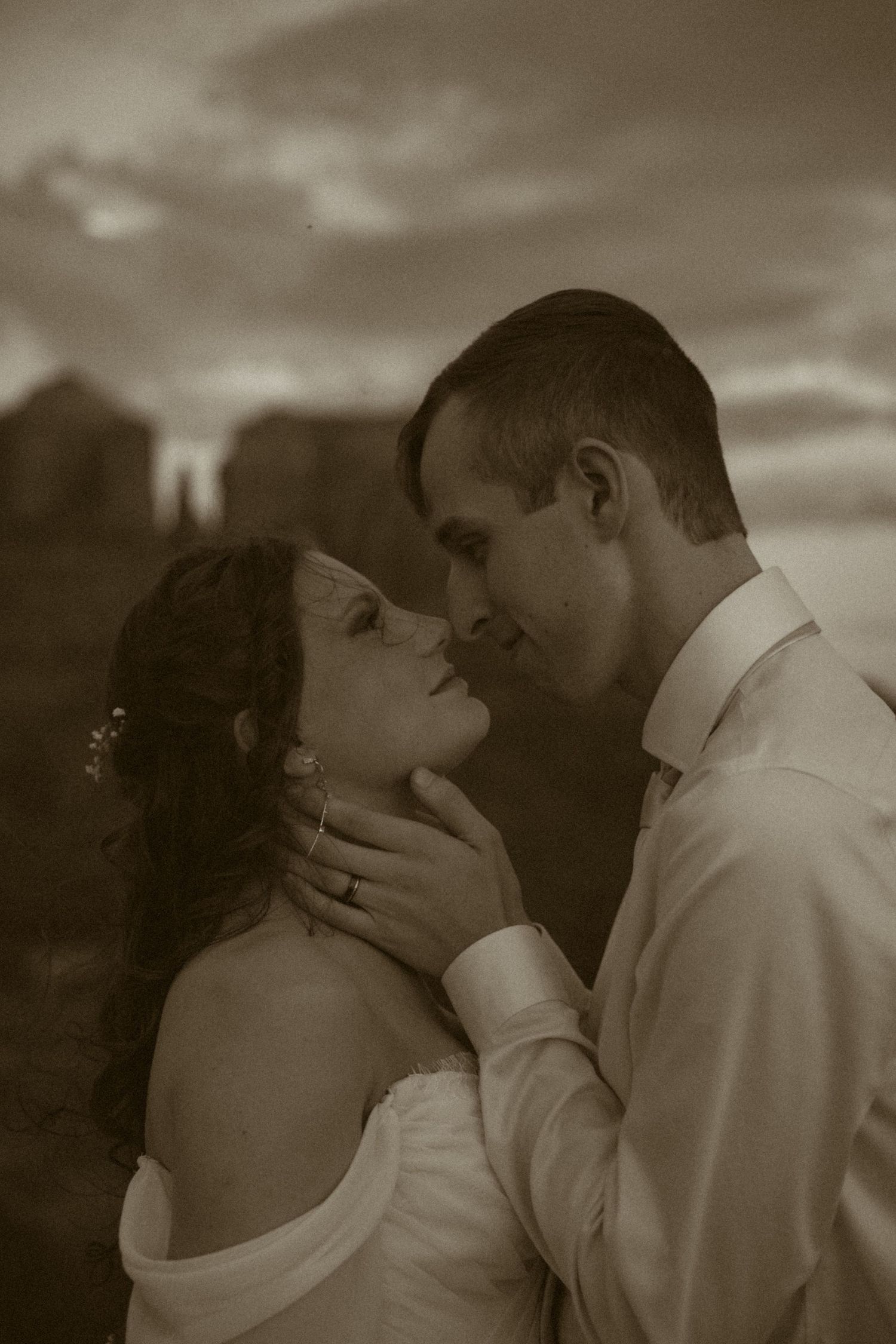 Sepia-toned romantic wedding photo of bride and groom nearly kissing at sunset with dramatic cloudy sky backdrop.