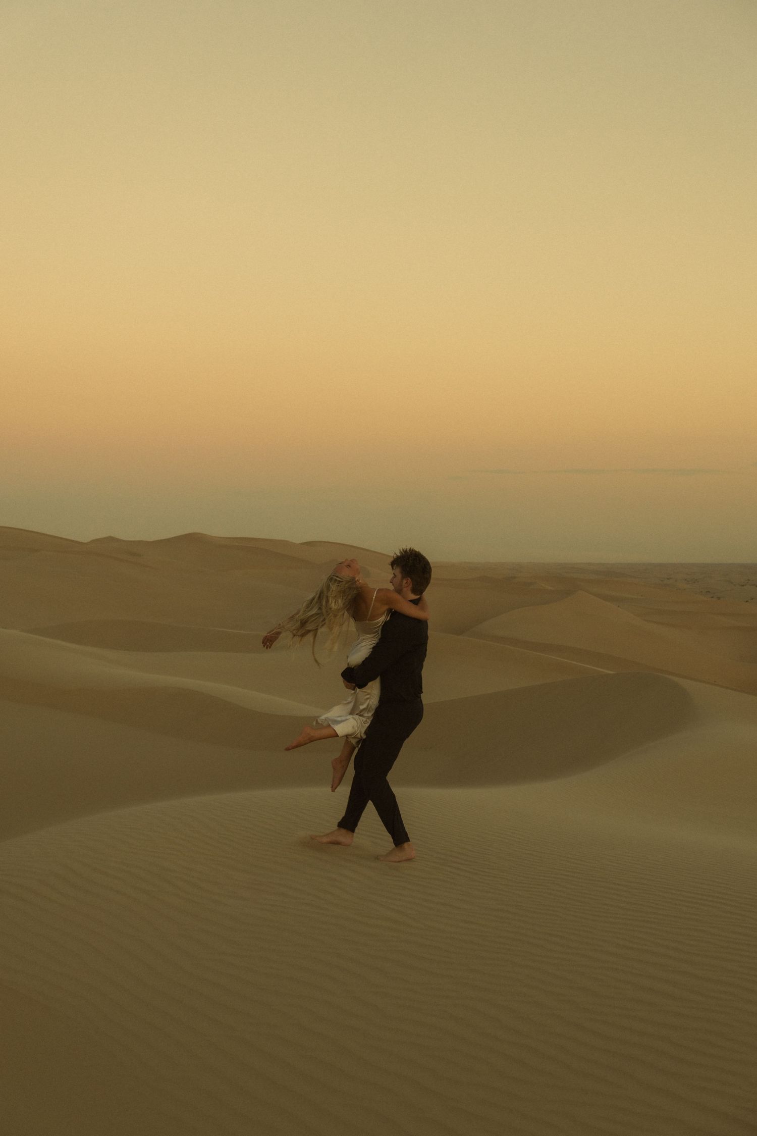 Couple dancing together on vast desert sand dunes during a warm golden hour sunset.