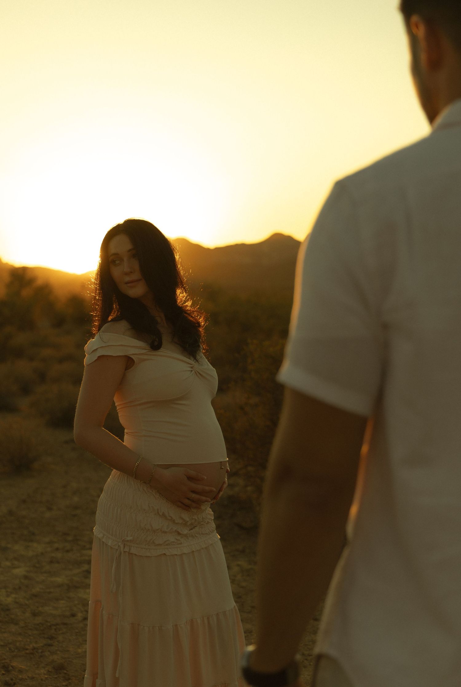 Pregnant woman in white dress cradles her baby bump at golden sunset in desert landscape with man in foreground.