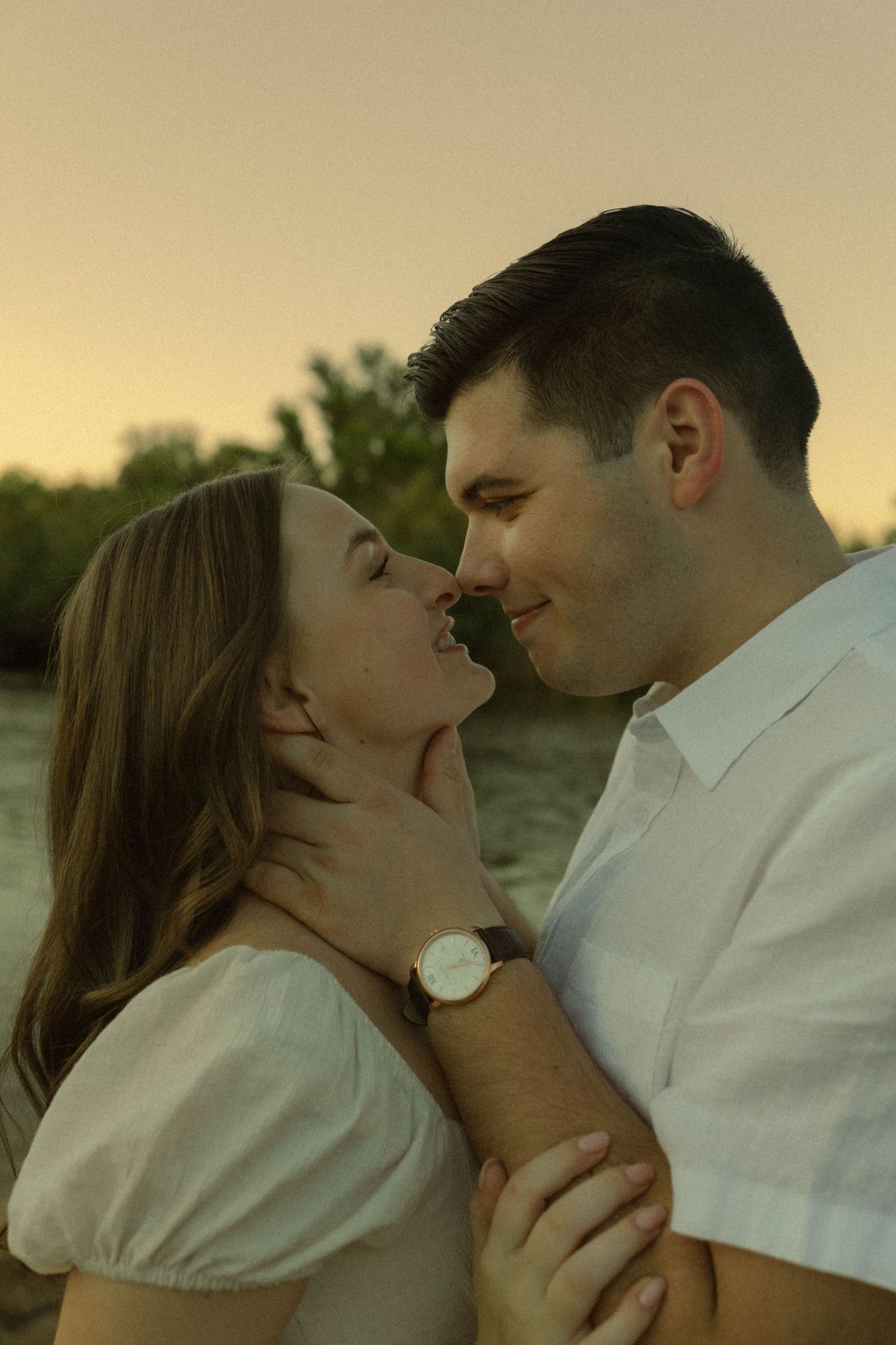 Romantic couple almost kissing at sunset, woman wearing white dress and watch, outdoors.
