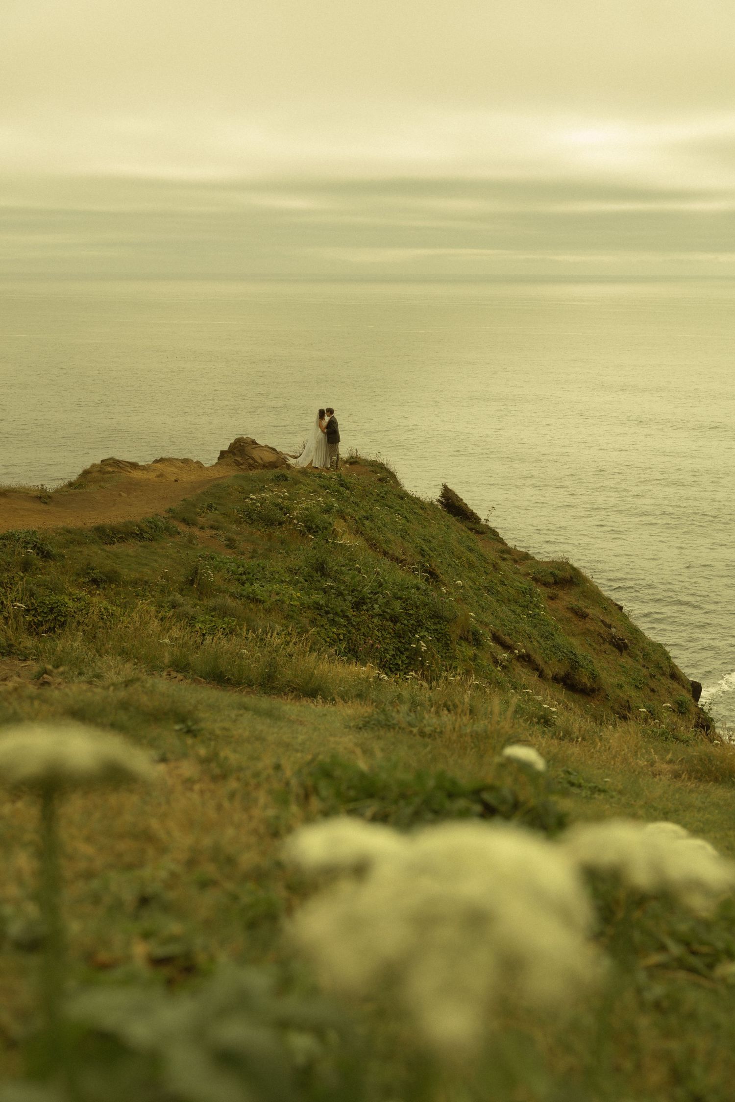 Couple standing on a coastal cliff overlooking the ocean, surrounded by wildflowers on a misty day.