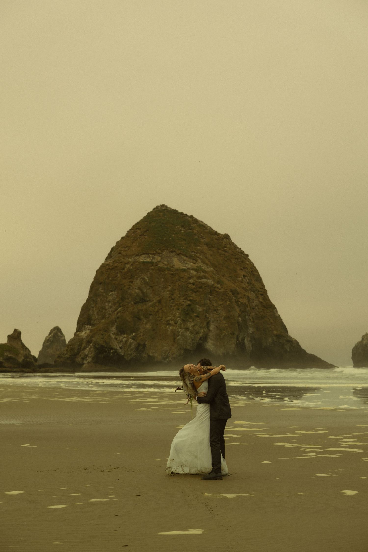 Wedding couple embraces and kisses on sandy beach with iconic Haystack Rock rising behind them.