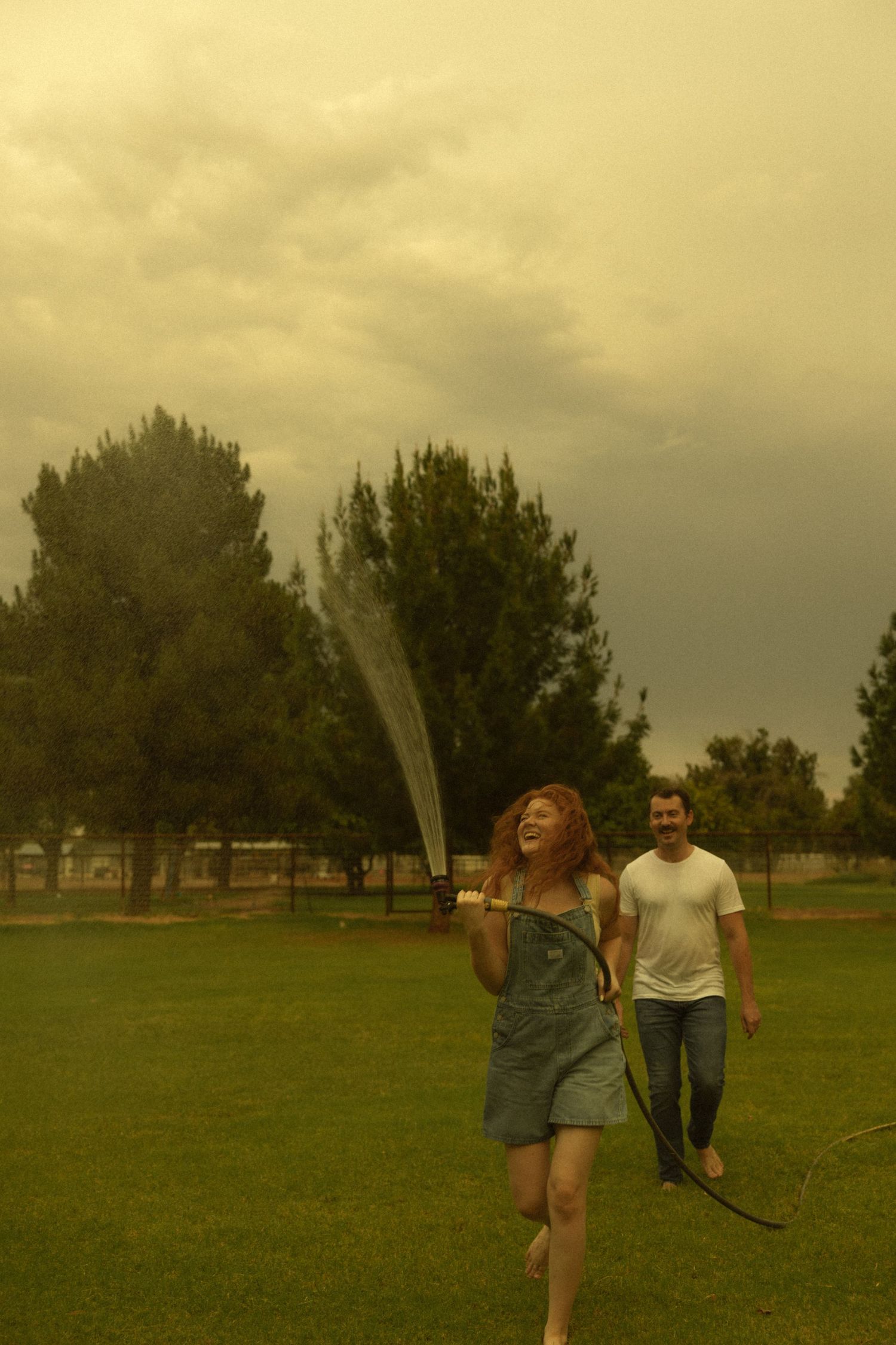 A laughing redhead woman in denim overalls sprays a hose outdoors while a man walks behind her on green grass.