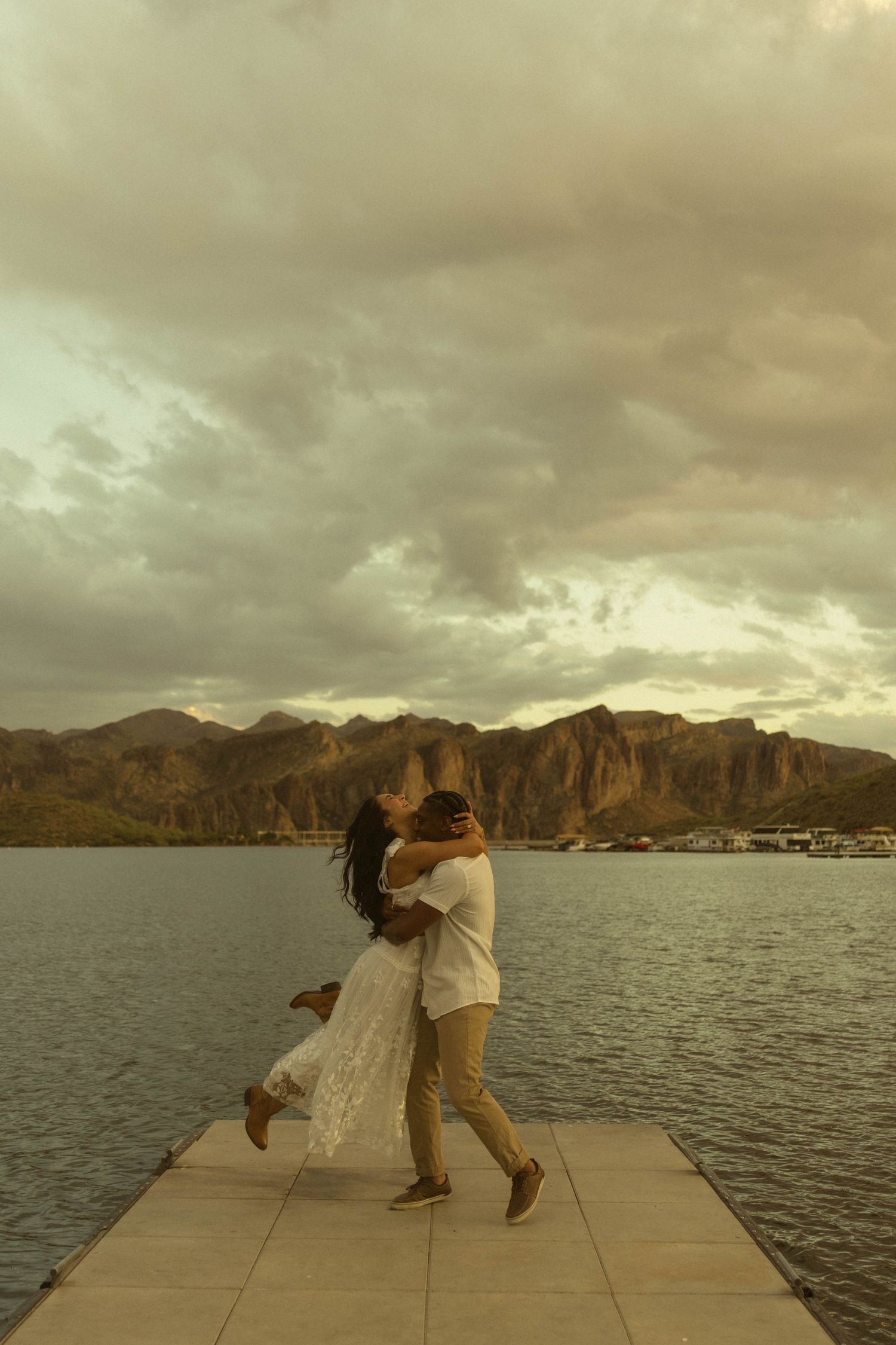 Couple embracing and kissing on a dock with mountains and cloudy sky over a lake in the background.
