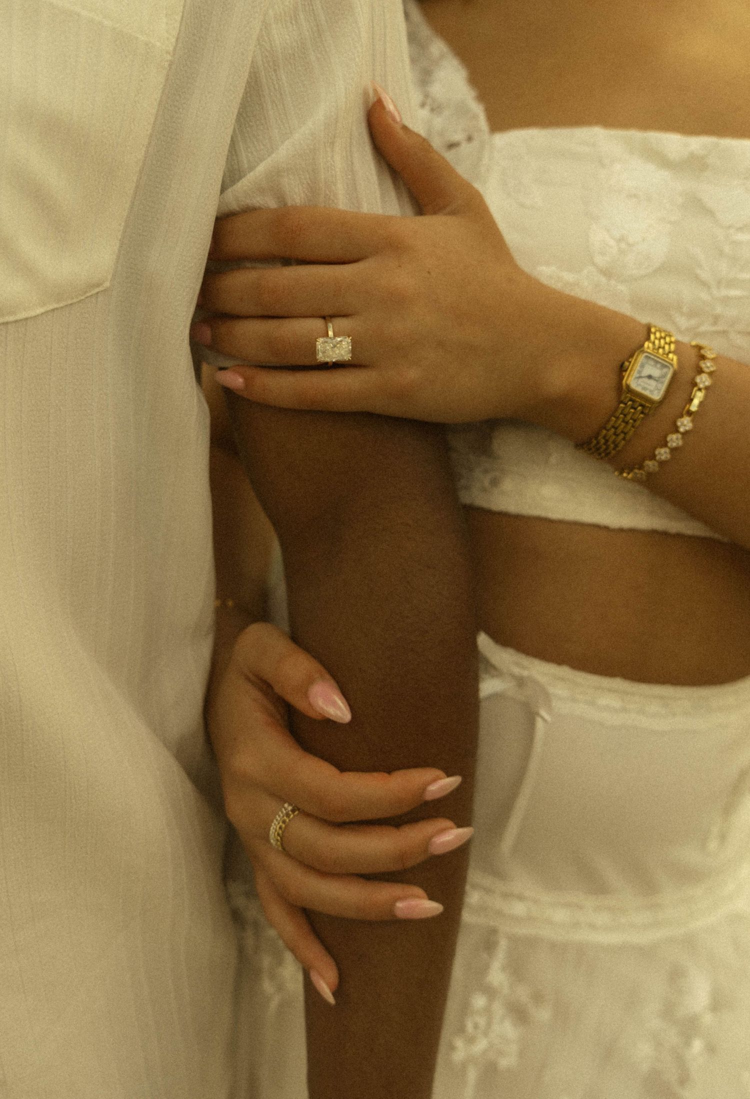 Close-up of two women's hands adorned with gold rings, a diamond engagement ring, and a gold watch, wearing white outfits.