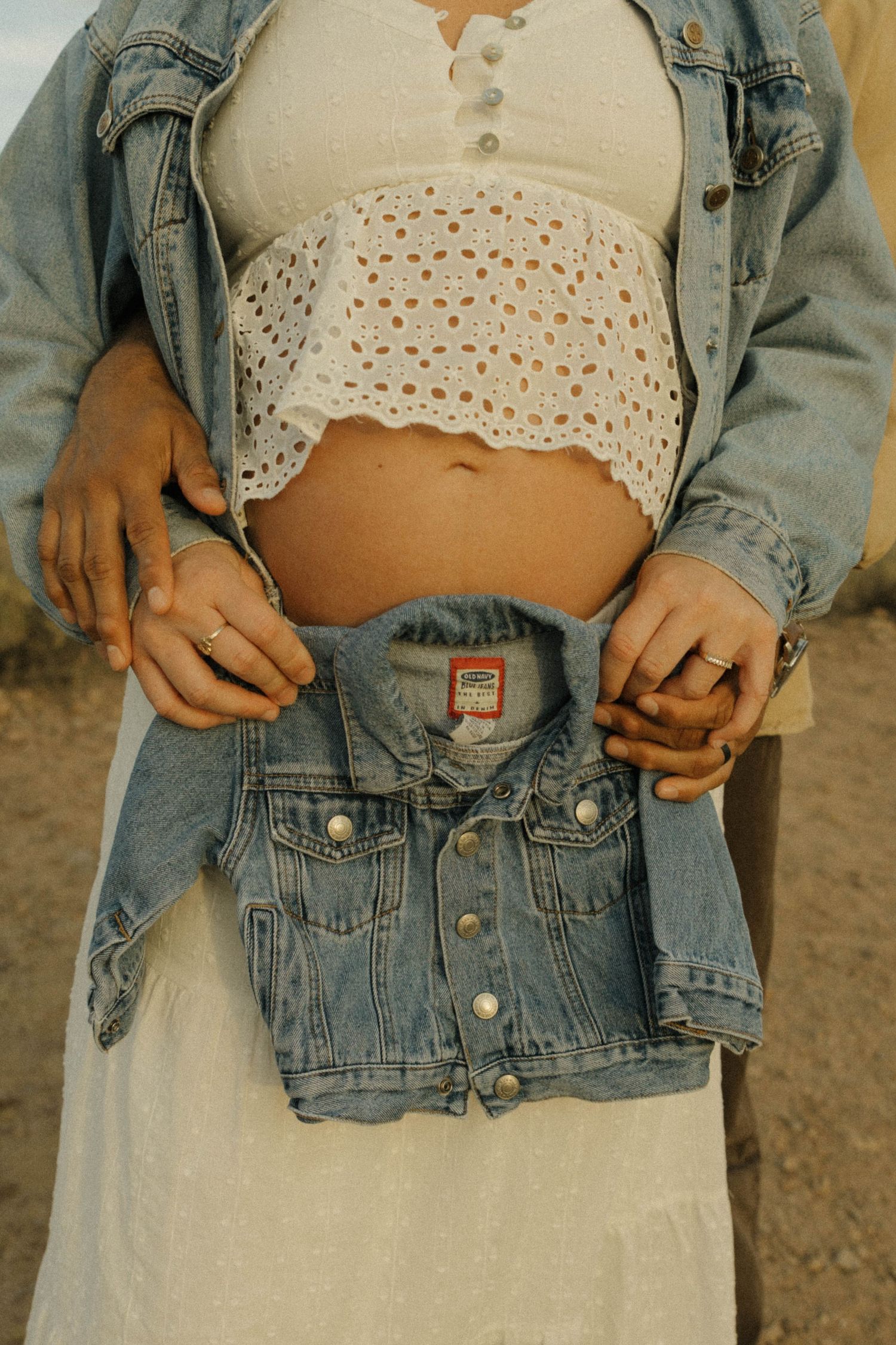 Pregnant woman holds tiny denim jacket over baby bump while partner embraces her outdoors.