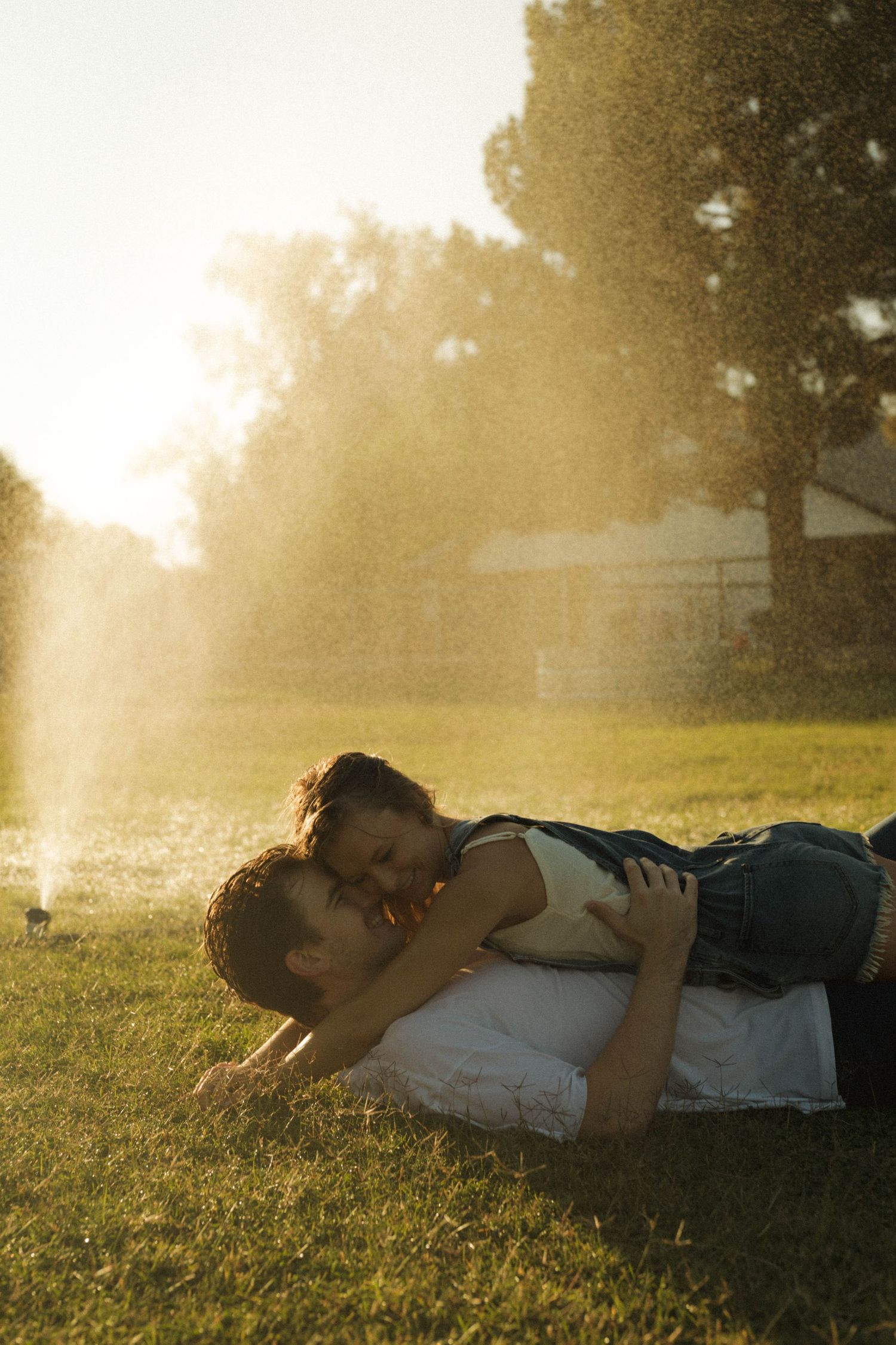 Couple sharing a romantic kiss lying on grass in golden sunset light with sprinklers misting in a lush backyard.