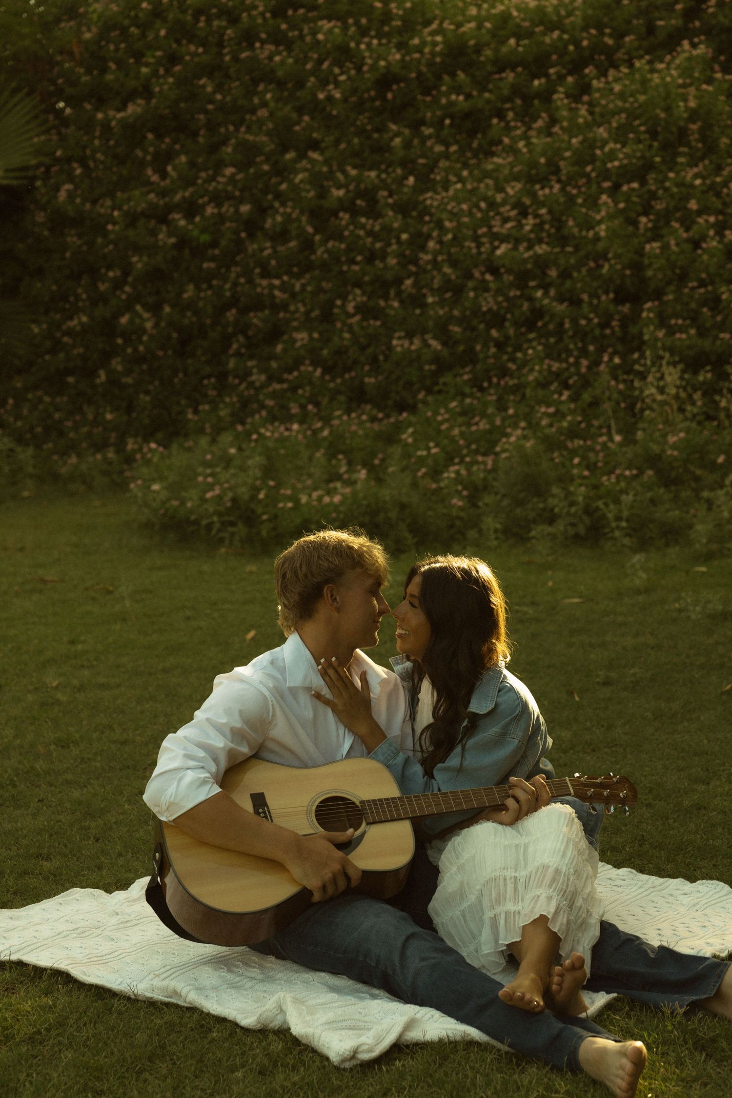 Romantic couple sitting on blanket in garden, man playing acoustic guitar while leaning in for a kiss with woman in white dress.