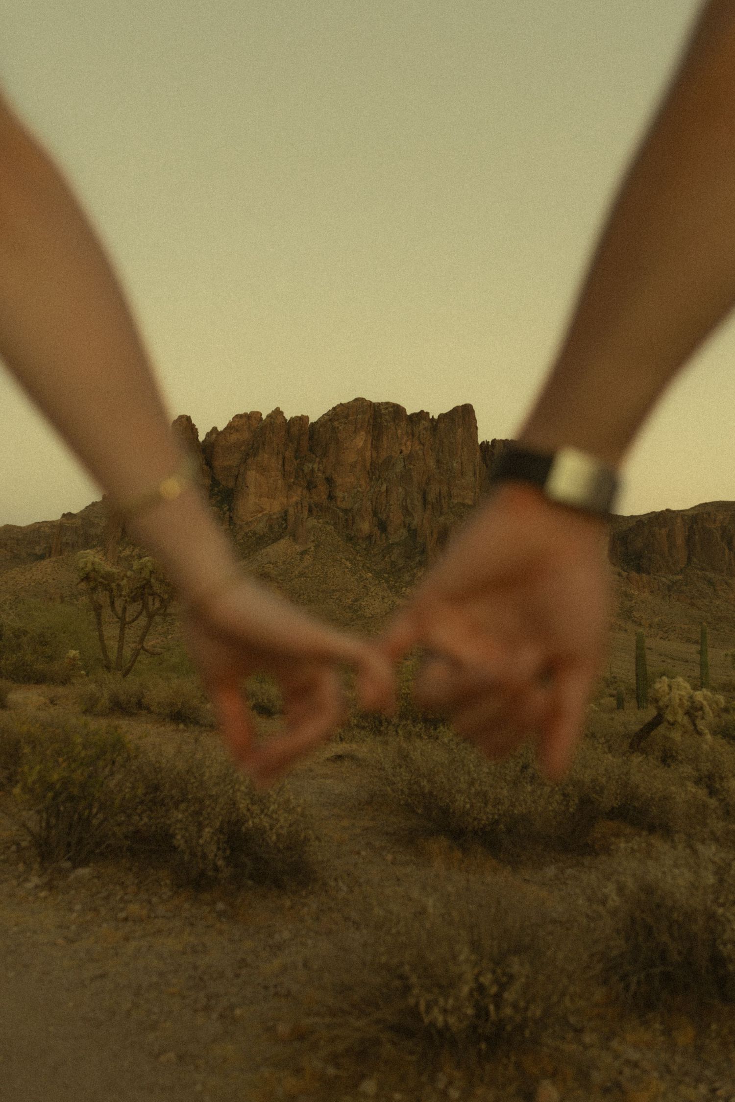 Couple lightly touching fingertips in blurred foreground with desert landscape and rocky butte at dusk.