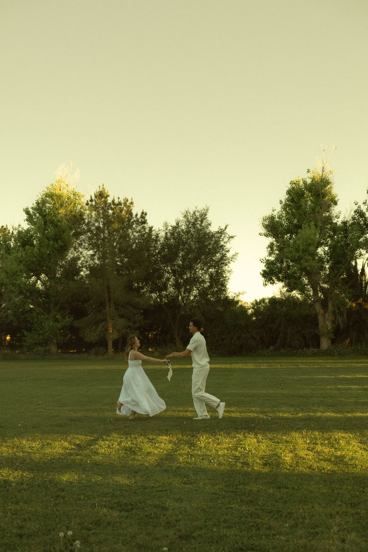 Expecting couple dancing joyfully on green grass field at sunset during outdoor maternity photoshoot.