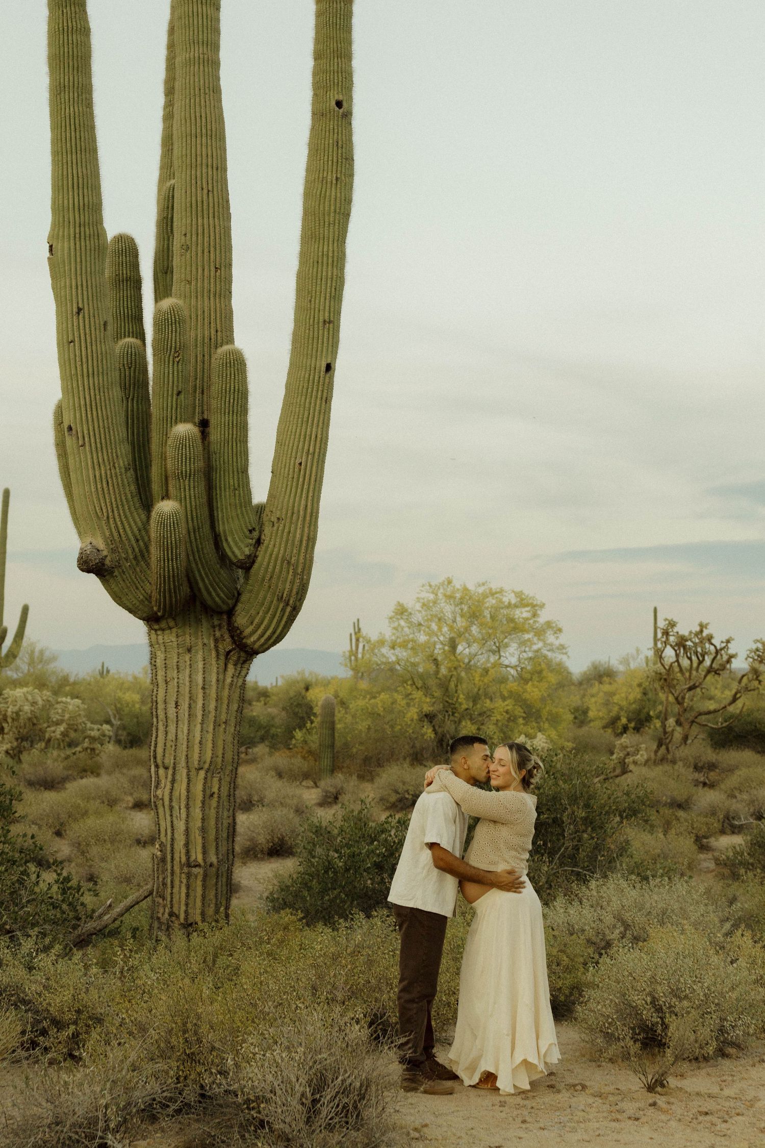 Couple embraces and kisses beside a large saguaro cactus in the Sonoran Desert.