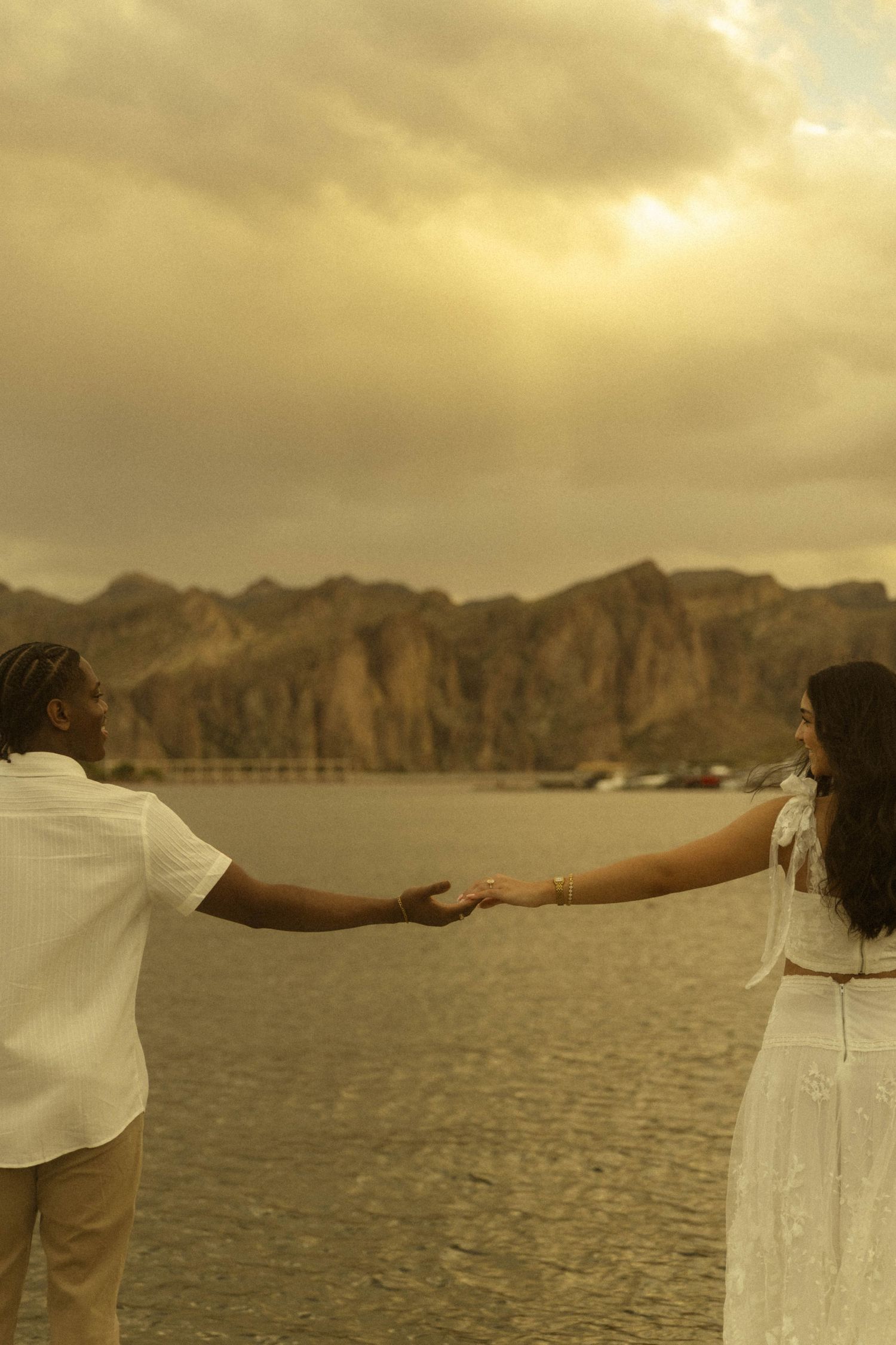 Couple holding hands by a serene lake with dramatic mountain backdrop and golden sunset sky.