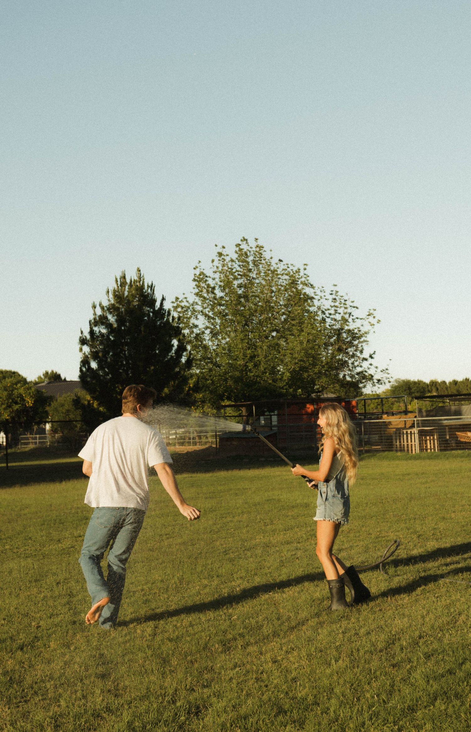 A girl sprays water from a hose at a man on a sunny green lawn with trees in the background.