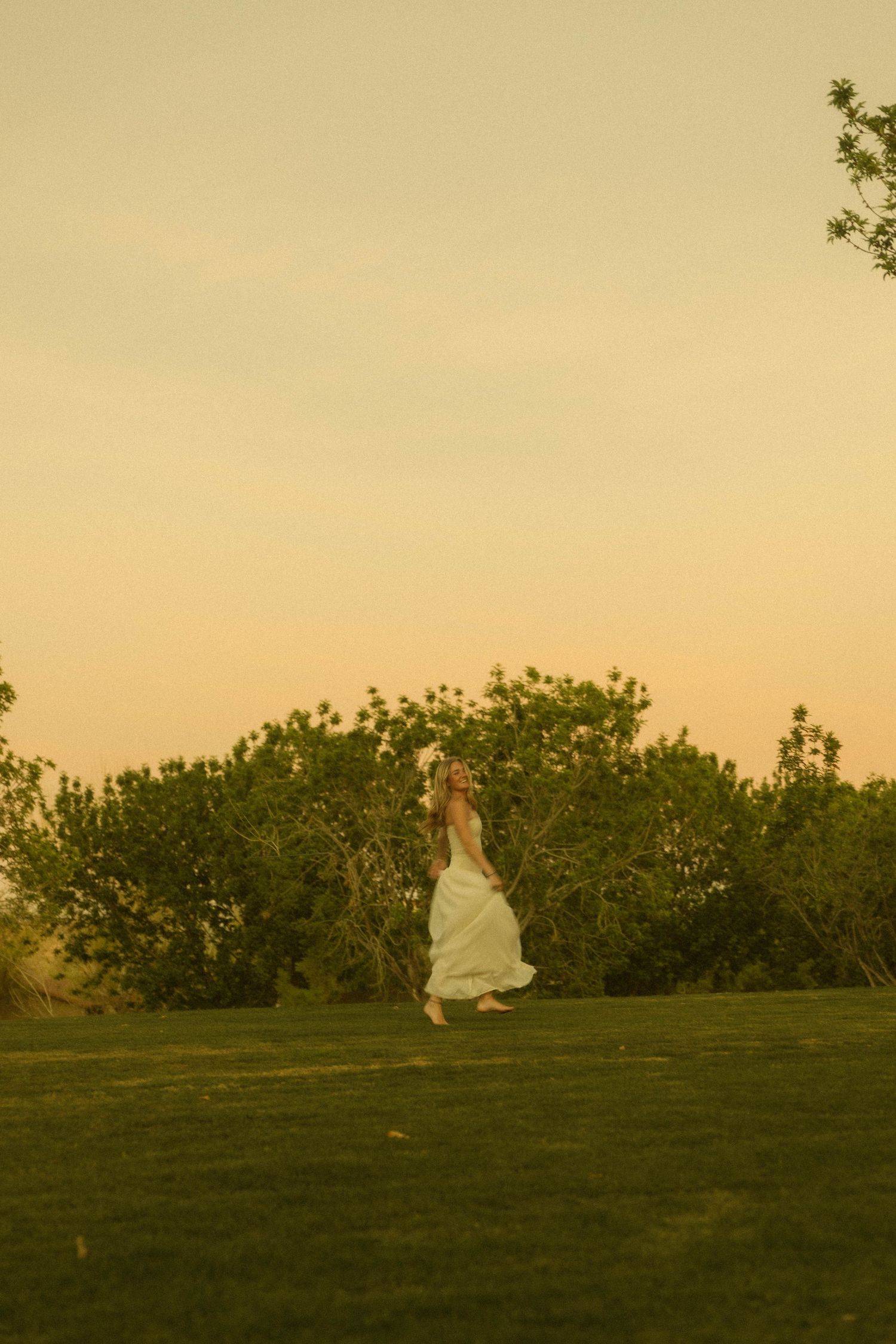 A woman in a flowing white dress walks gracefully across a lush green lawn at golden hour sunset.