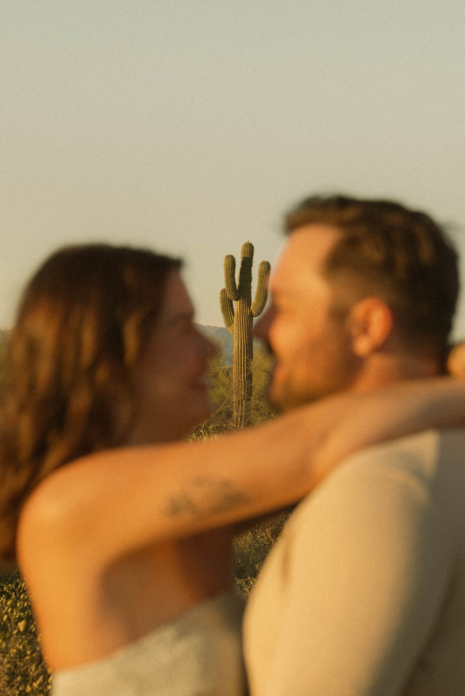 Couple sharing a romantic kiss at golden hour with a saguaro cactus in the desert background.