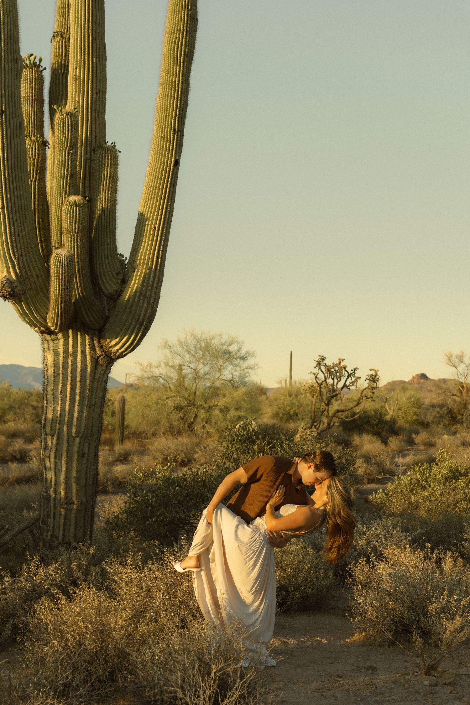 Couple sharing a romantic dip kiss in the Arizona desert surrounded by tall saguaro cacti at golden hour.