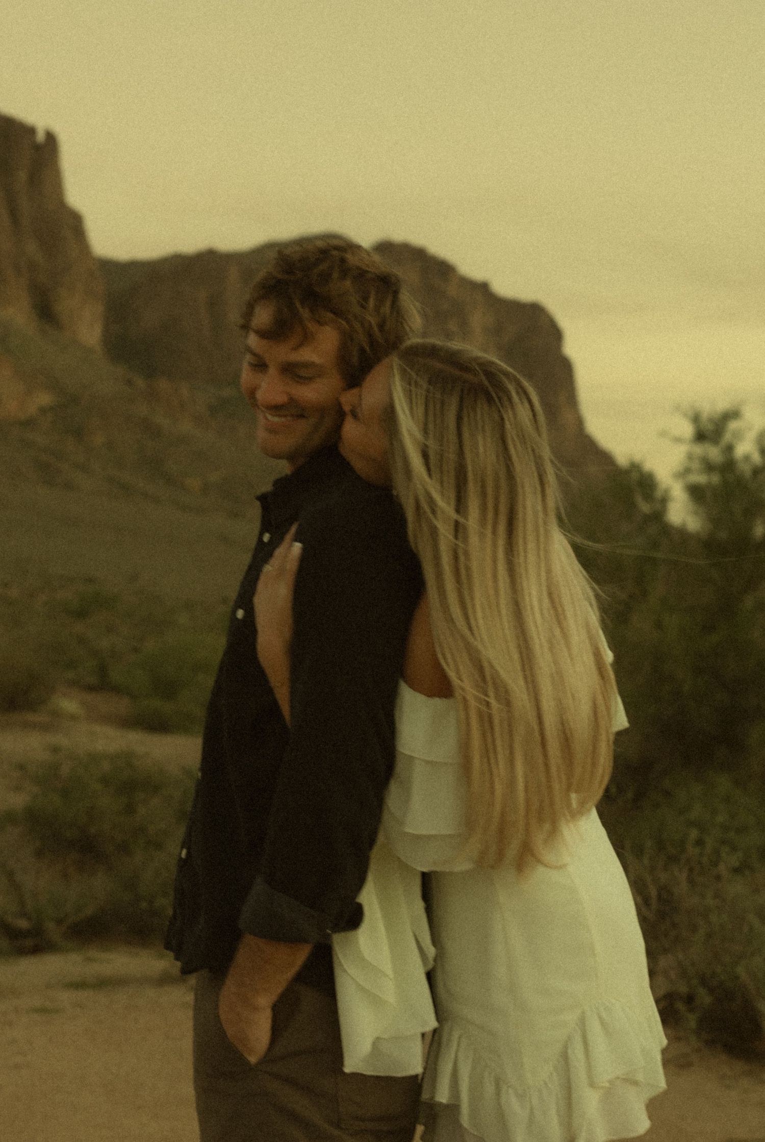 Smiling man in a dark shirt embraced from behind by a blonde woman in a white dress, with desert mountains and hazy sky behind them.
