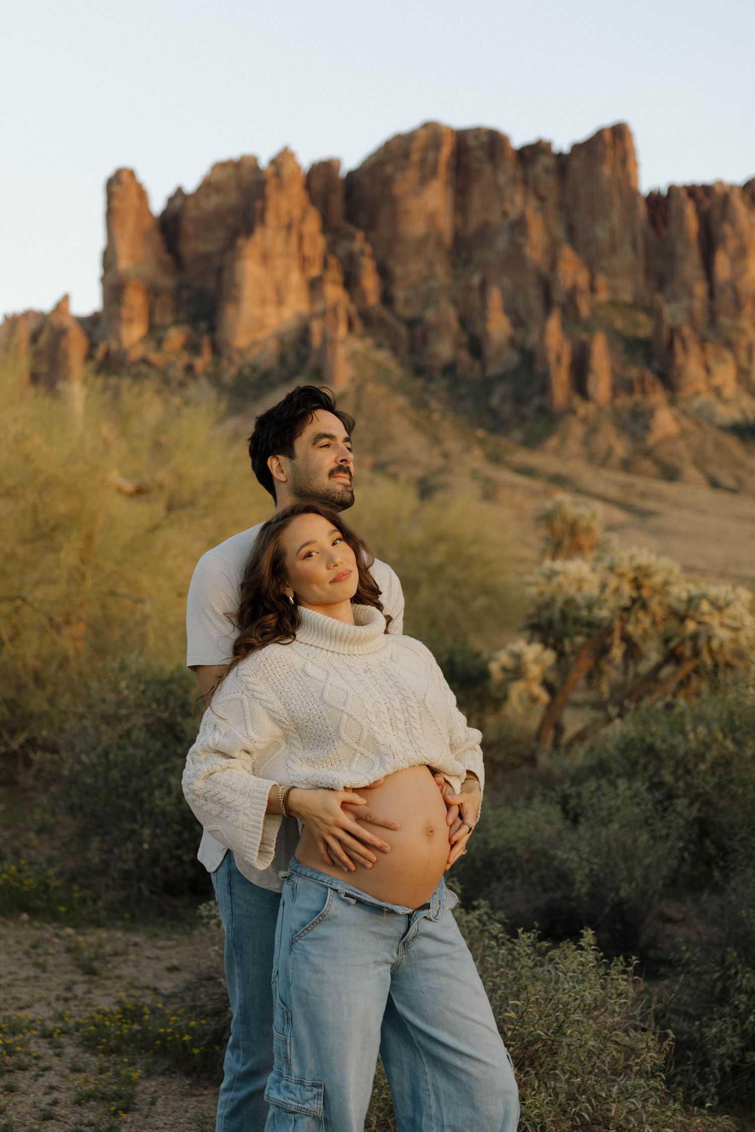 A maternity photoshoot in front of majestic desert cliffs and mountains during golden hour.