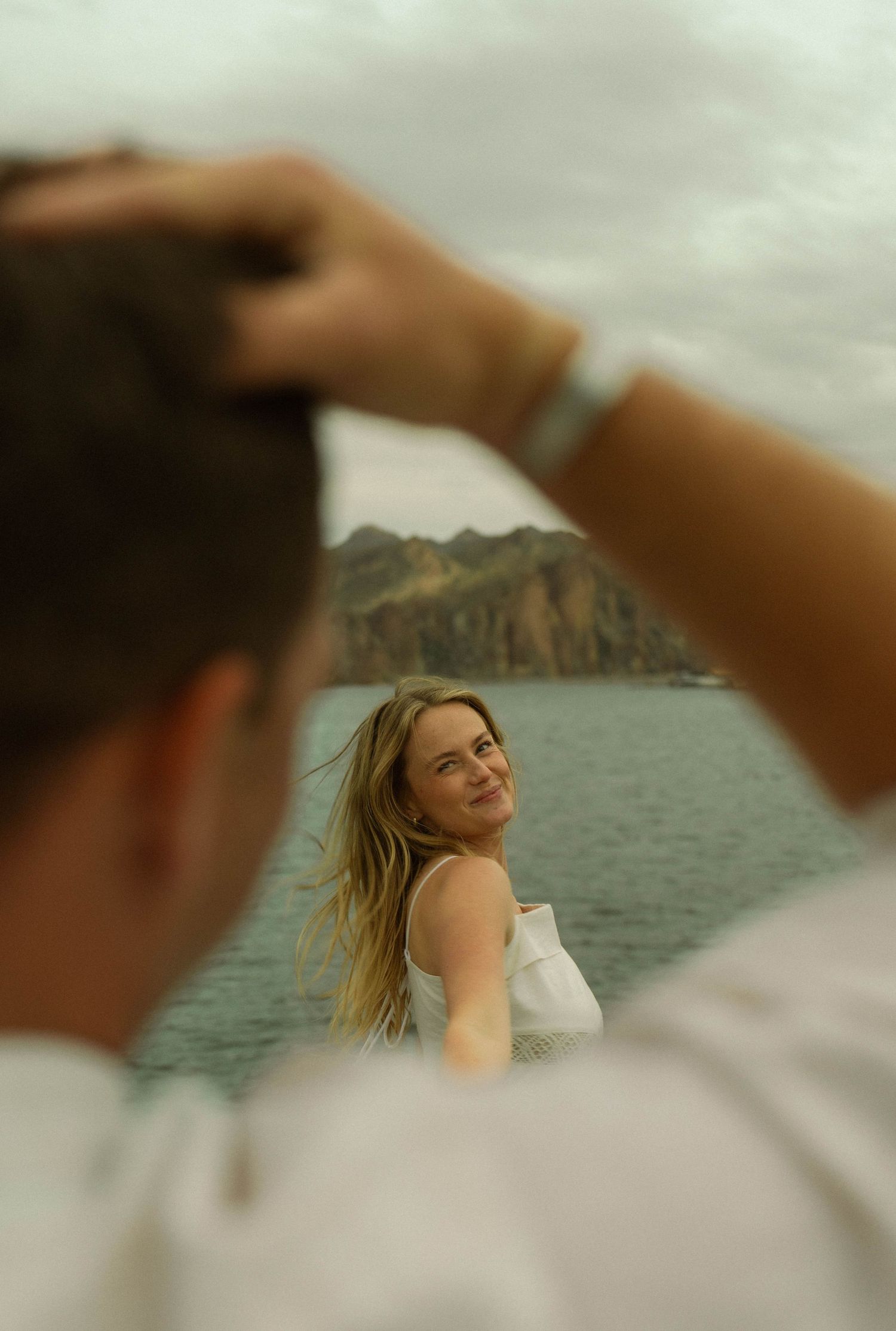 A dreamy seaside portrait with mountains in the background and flowing blonde hair catching the wind.
