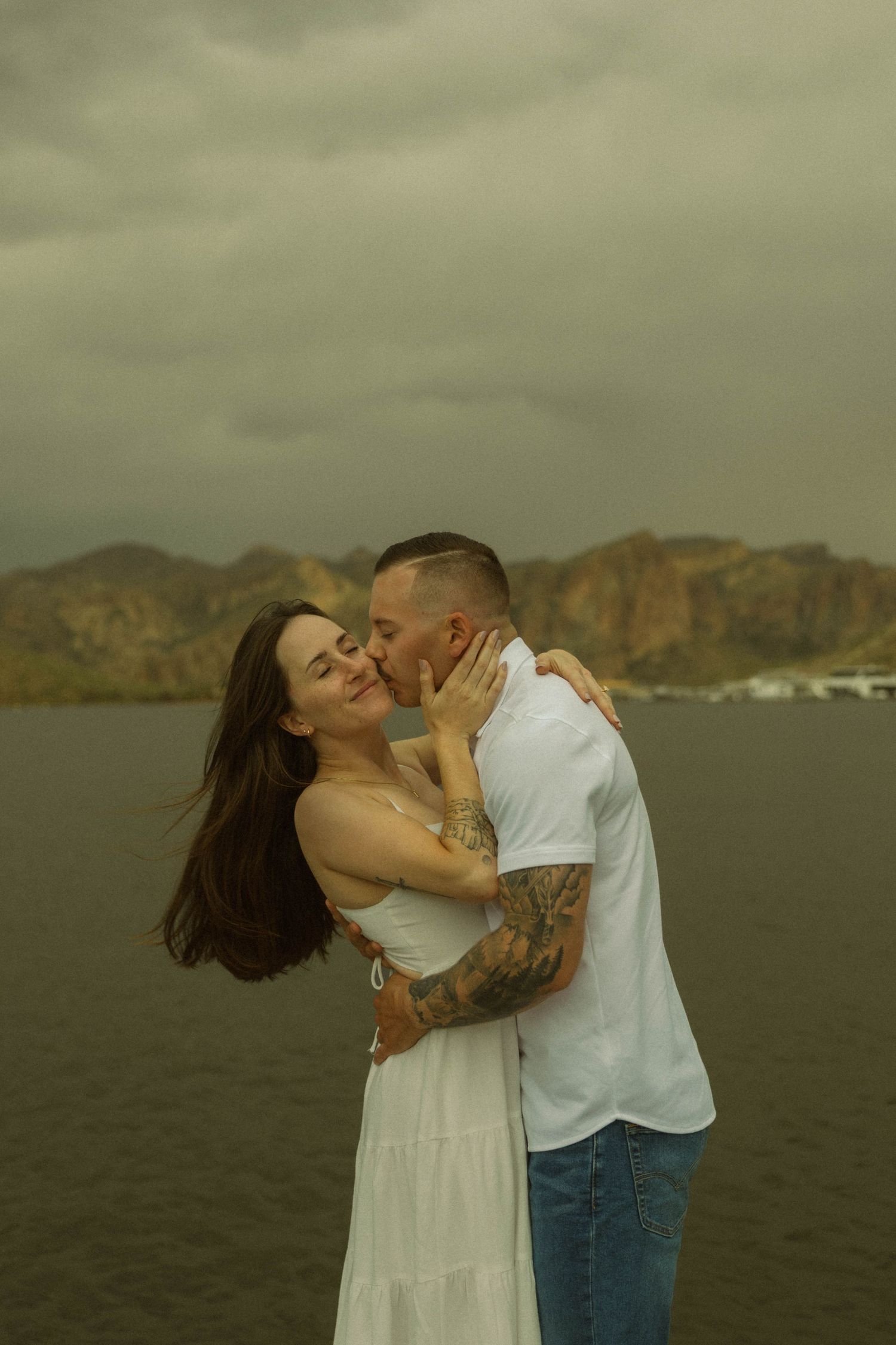 Loving couple embracing by a lake with dramatic mountains in the background.