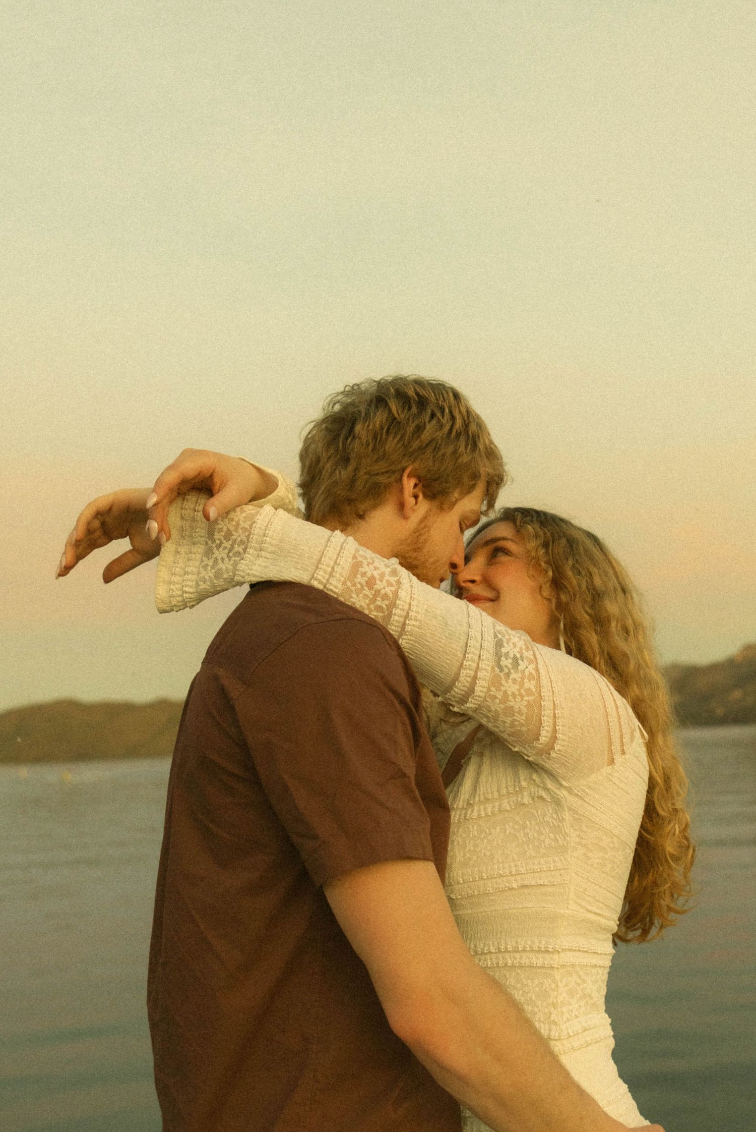 A tender embrace between a couple at golden hour with mountains and water in the background.