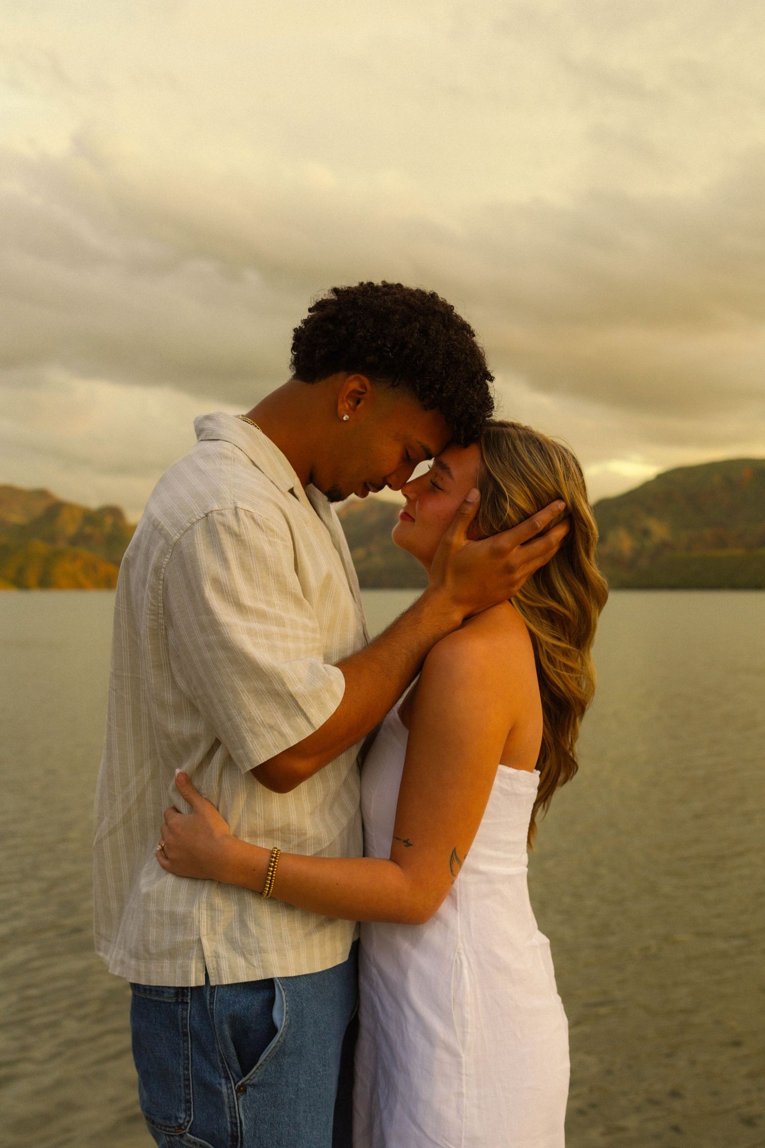 A romantic couple embraces against a sunset lake backdrop with mountains in the distance.