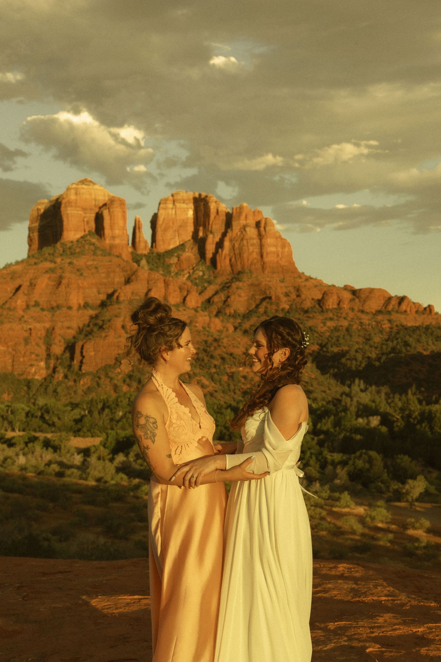 Two brides in elegant gowns embrace at sunset with Cathedral Rock in Sedona, Arizona glowing behind them.