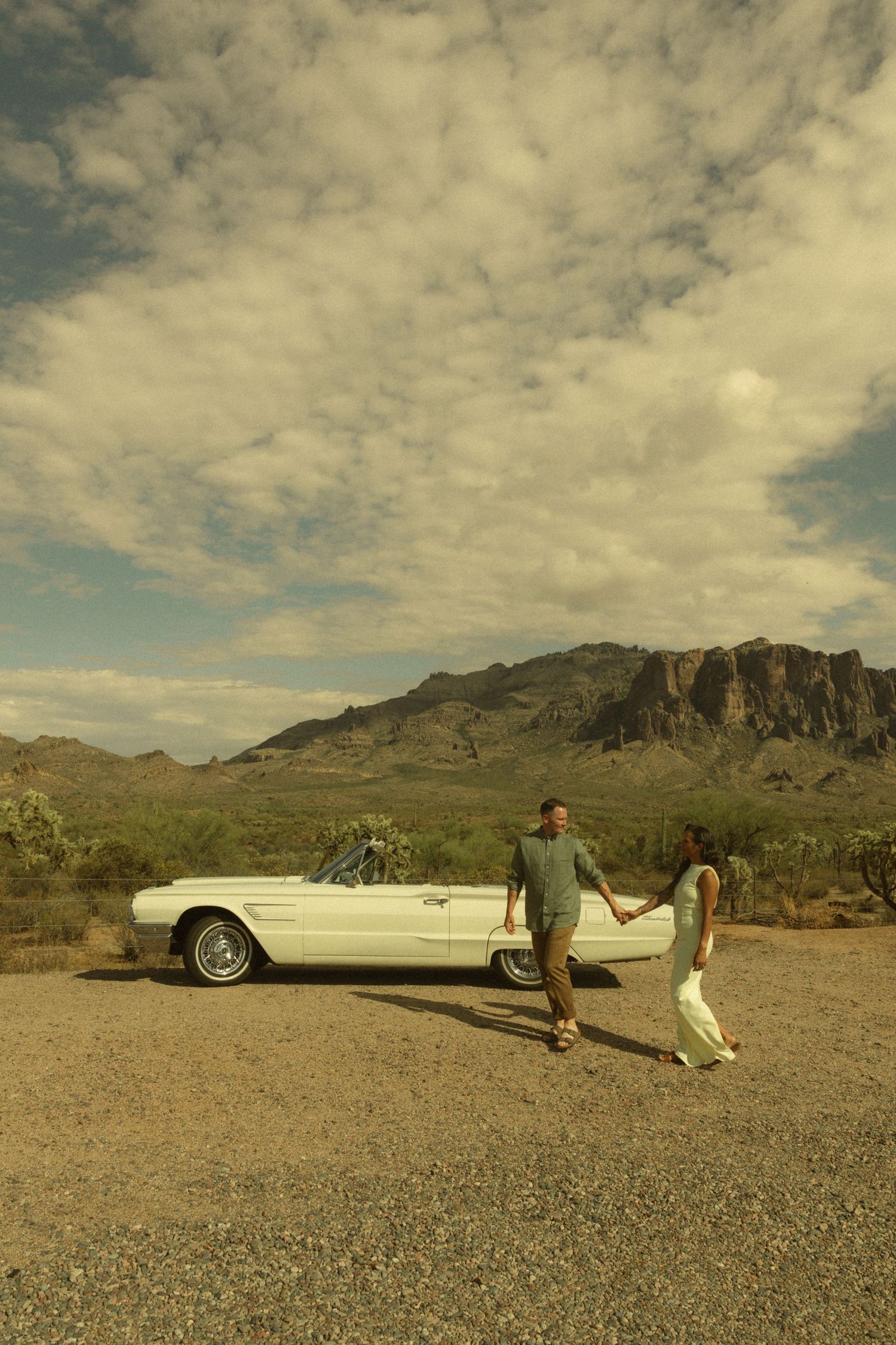 Couple holding hands by a white vintage convertible in a desert landscape with mountains and dramatic clouds.