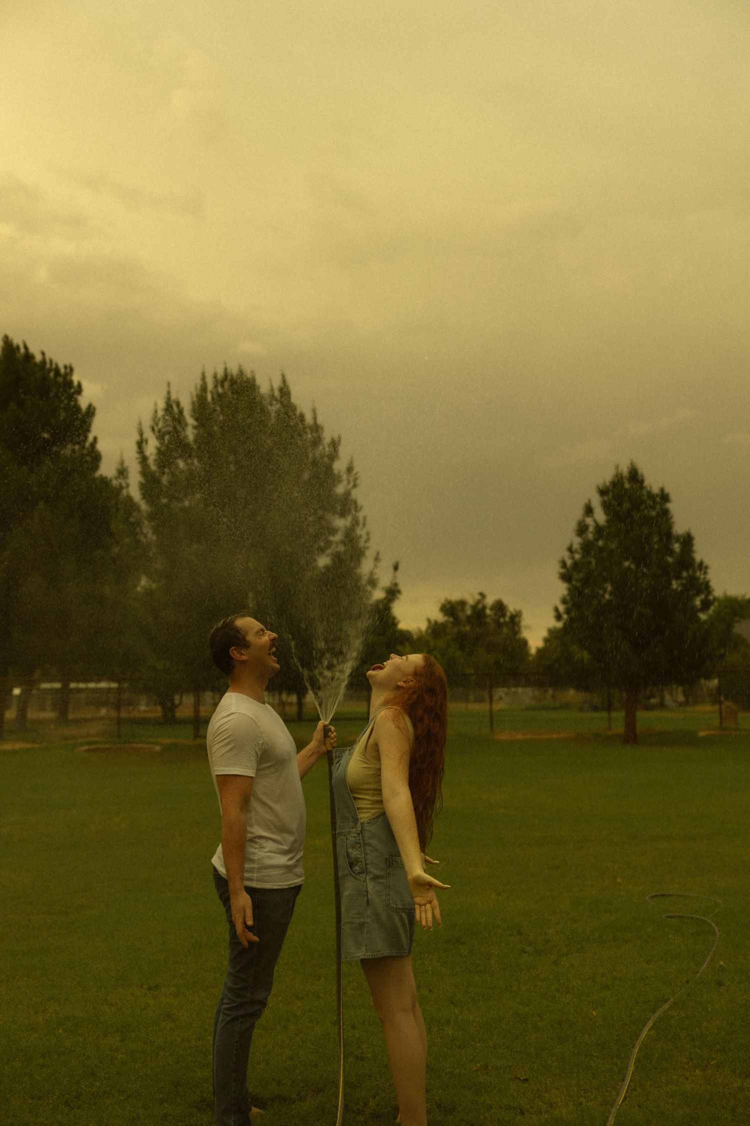 Couple standing together in park as man sprays water hose upward under moody golden stormy sky.