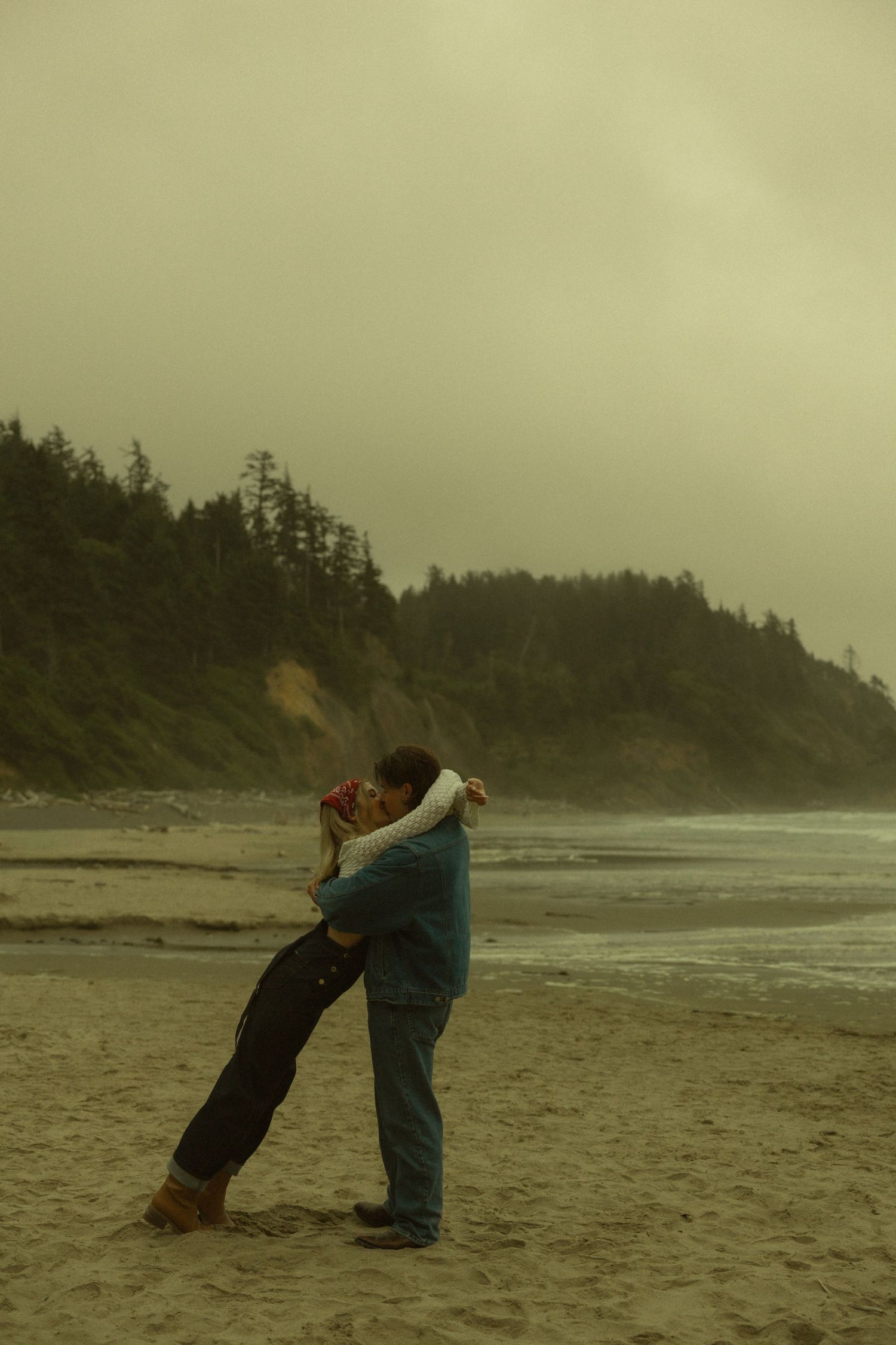 Romantic couple embracing on a foggy Pacific Northwest beach with evergreen cliffs in the background.