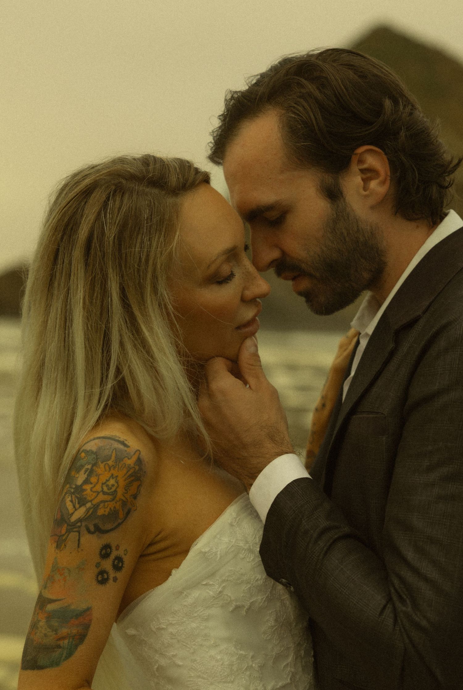 Tattooed bride and groom share an intimate moment at beach wedding, foreheads nearly touching.