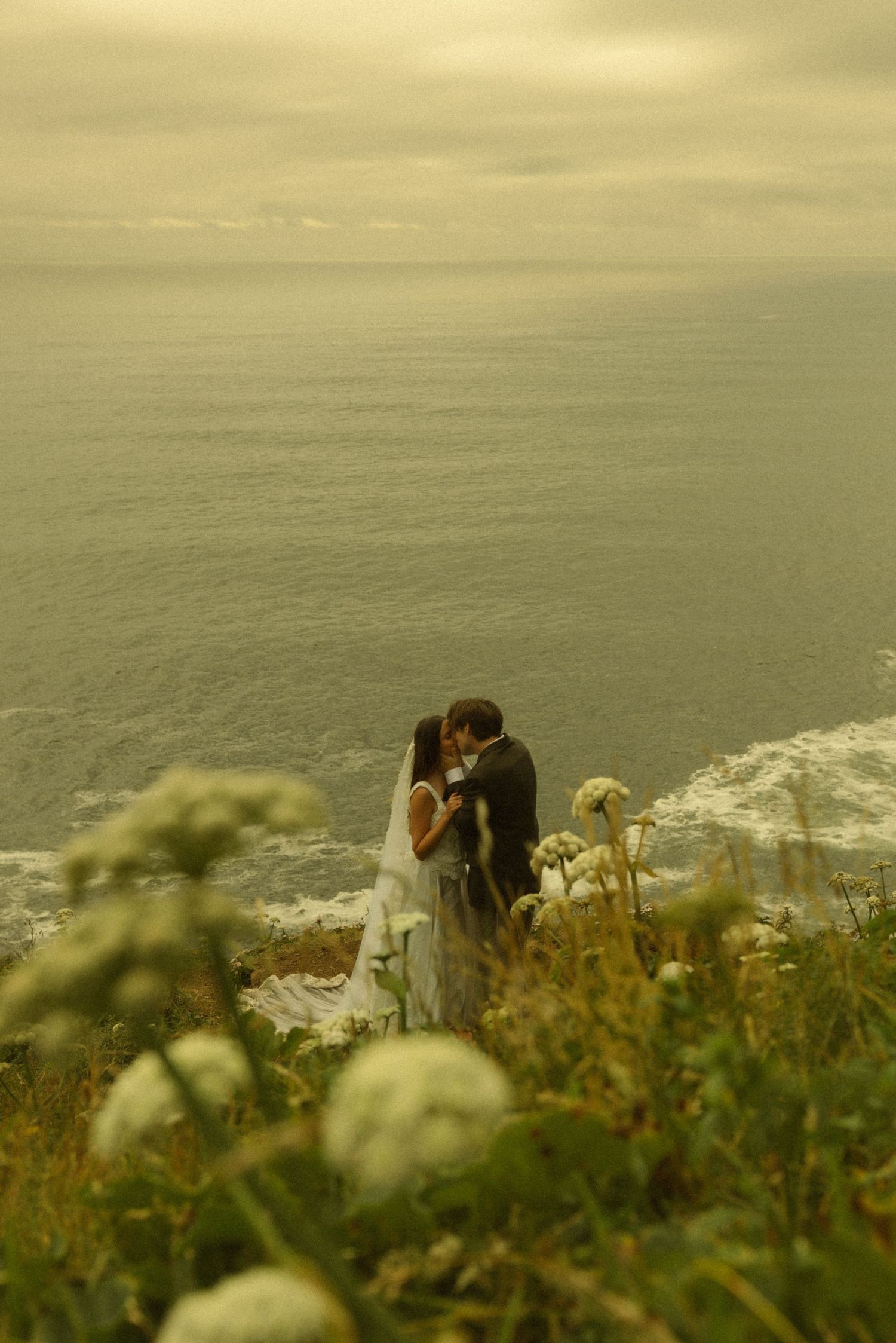 A couple shares a romantic kiss on a coastal cliff overlooking the ocean, surrounded by wildflowers on their wedding day.