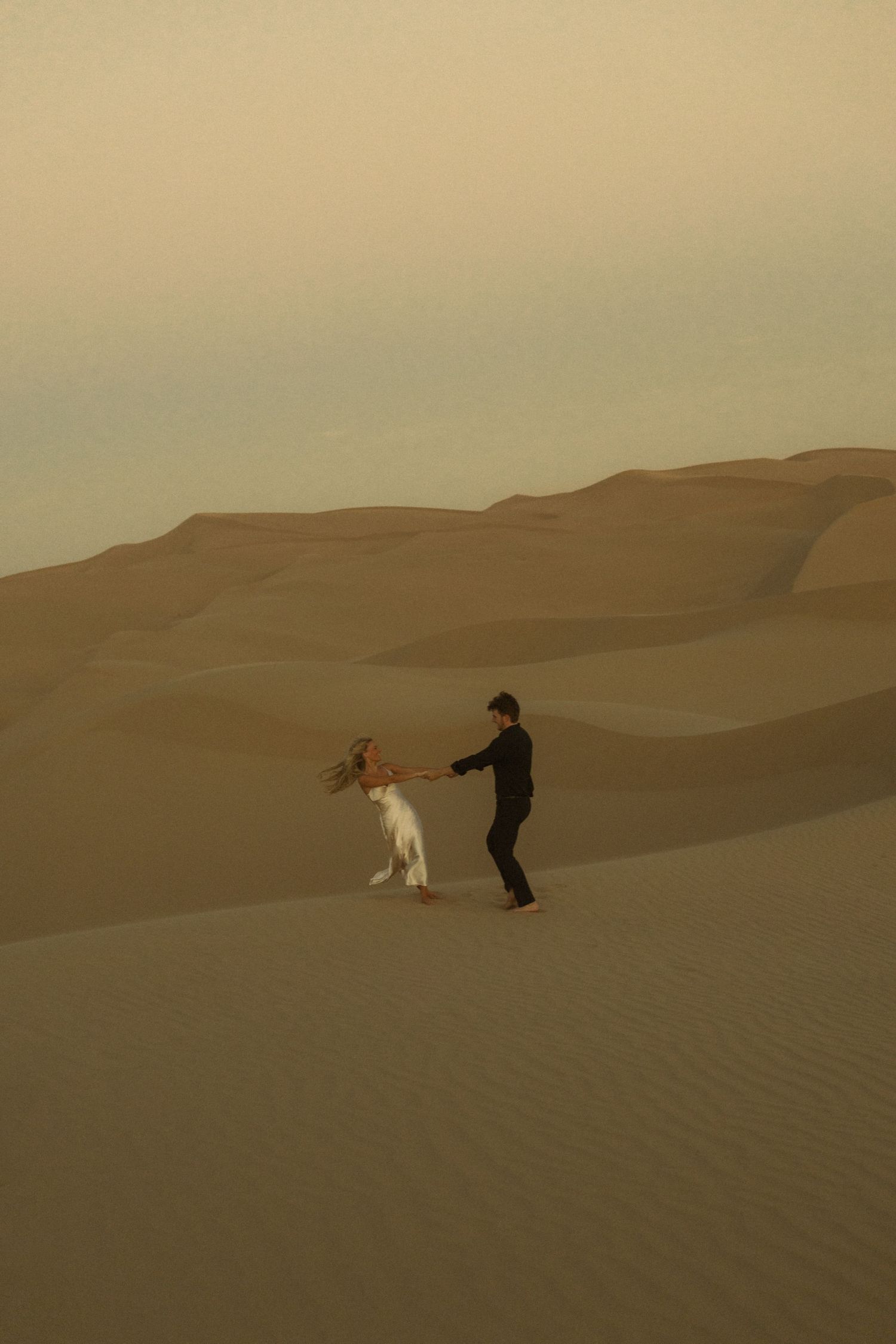 Couple dancing together on vast sandy desert dunes under a hazy sky at dusk.